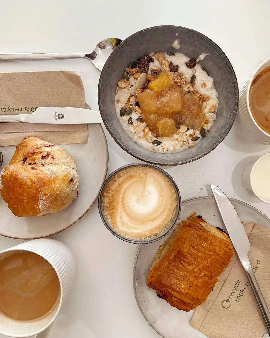 Aerial view of a breakfast spread featuring creamy porridge with fruit and nuts, a flaky pastry, a scone, and two cups of coffee, conveying a cozy morning vibe.