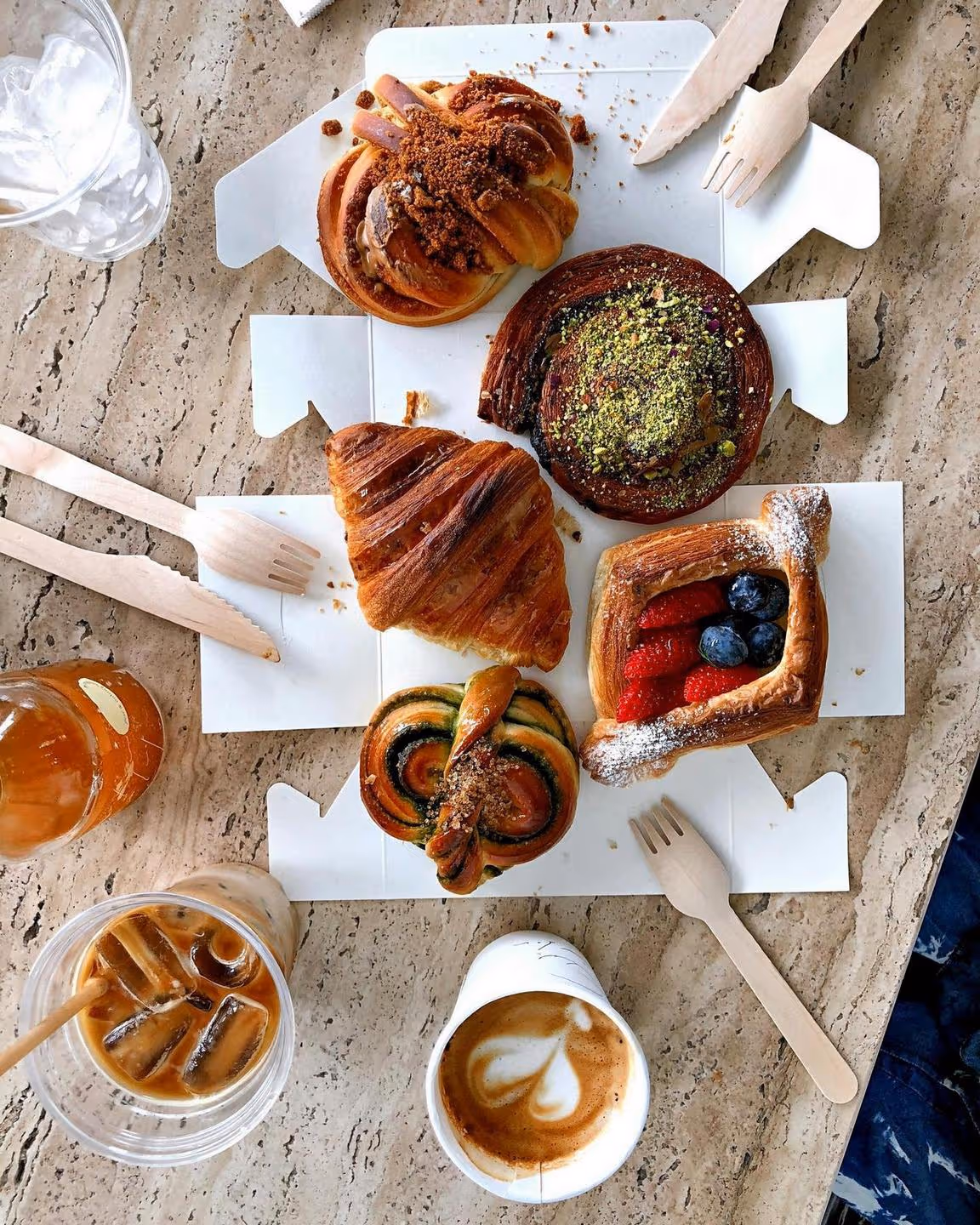 Assorted pastries with berries, pistachio, and cinnamon swirls on a table, surrounded by iced coffee, cappuccino, wooden forks, and a refreshing, inviting vibe.