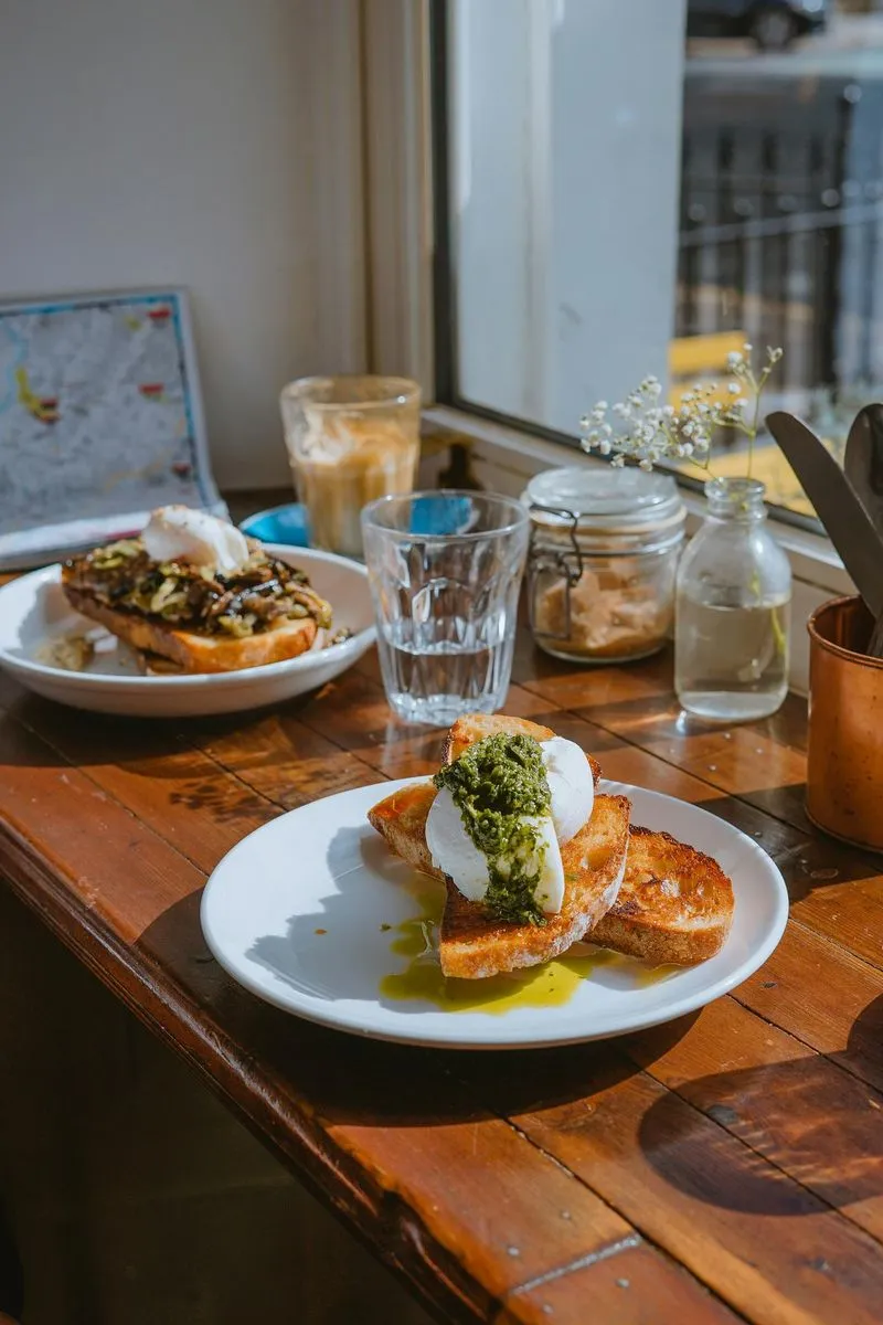 Plates of toast topped with poached eggs and pesto sit on a wooden table by a sunny window. Nearby are a latte, glass, and small vase. Cozy brunch setting.