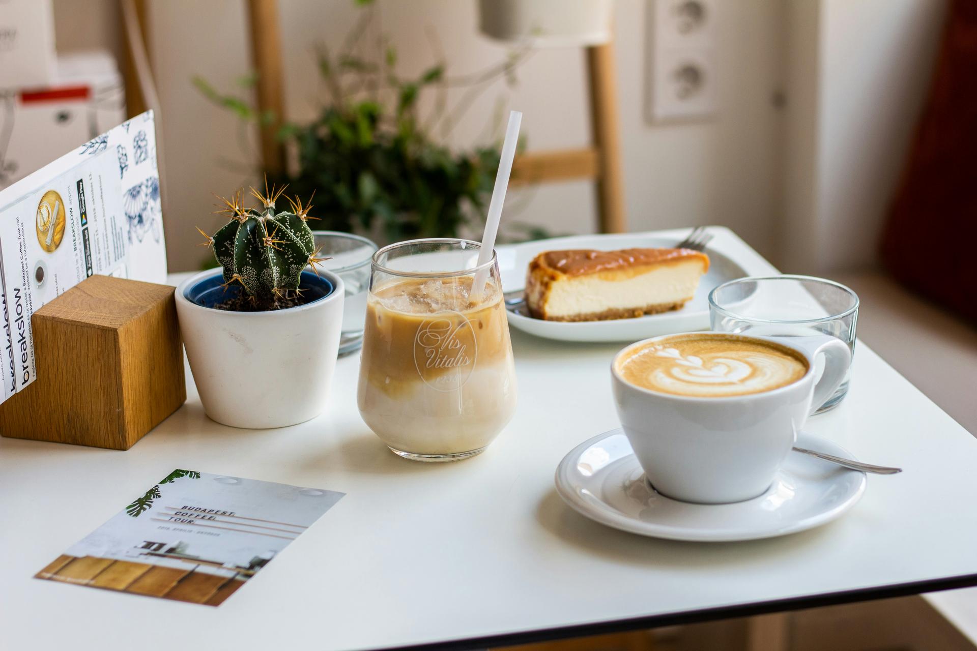 A cozy café table with a cappuccino, iced coffee, and a slice of cheesecake. A small potted cactus adds charm, and soft light creates a relaxing mood.