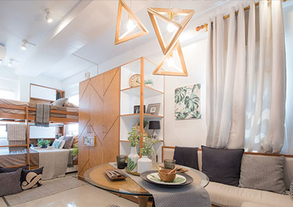 Modern living space with a beige sofa, black and gray cushions, round glass coffee table, wooden shelves, bunk beds, and triangular pendant lights.