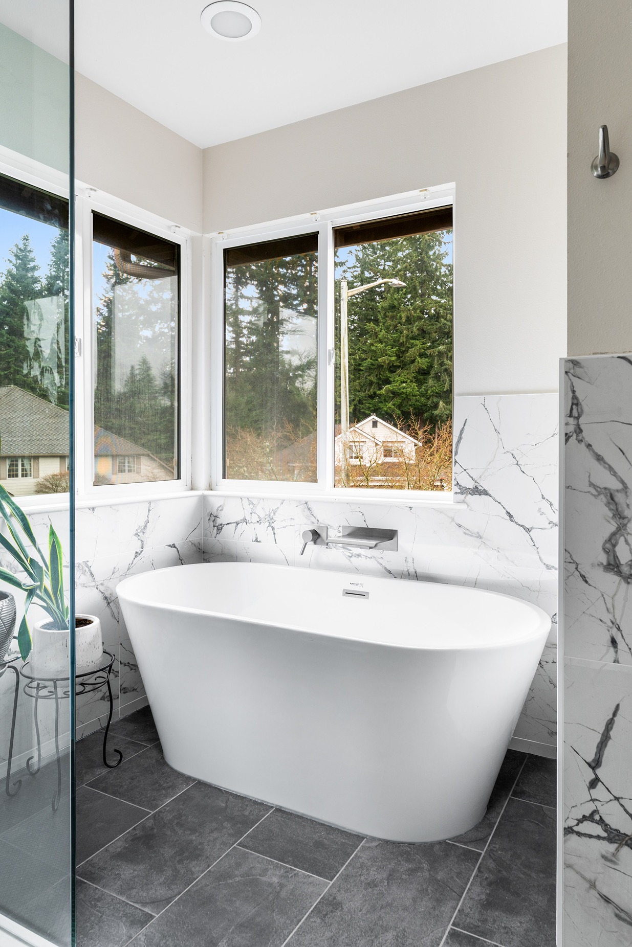 Bright modern bathroom corner with a freestanding white bathtub, marble wall tiles, large windows, and a potted plant beside the tub.