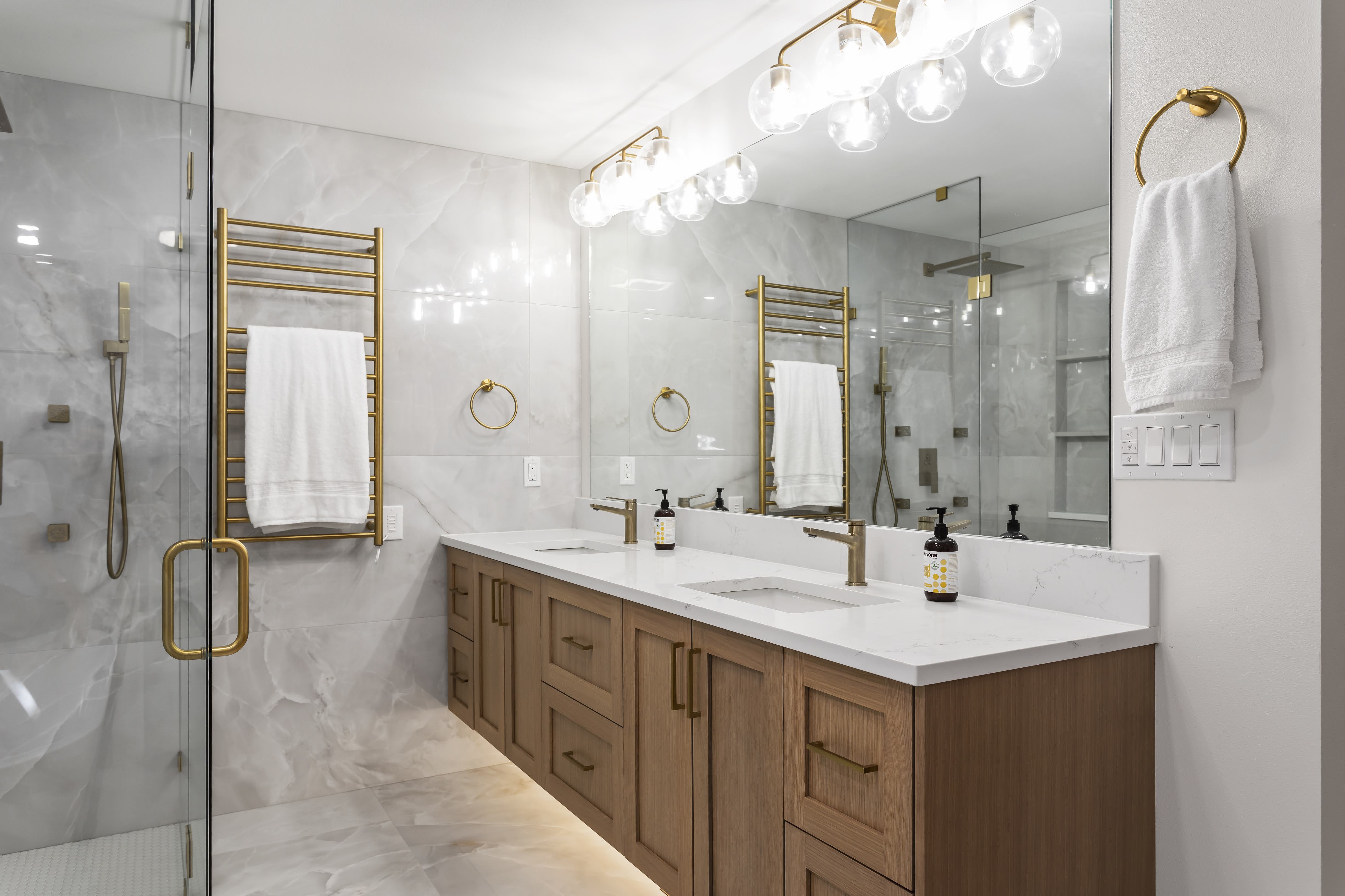Elegant bathroom design with wood cabinetry, gold fixtures, large illuminated mirror, and a walk-in shower with gold accents