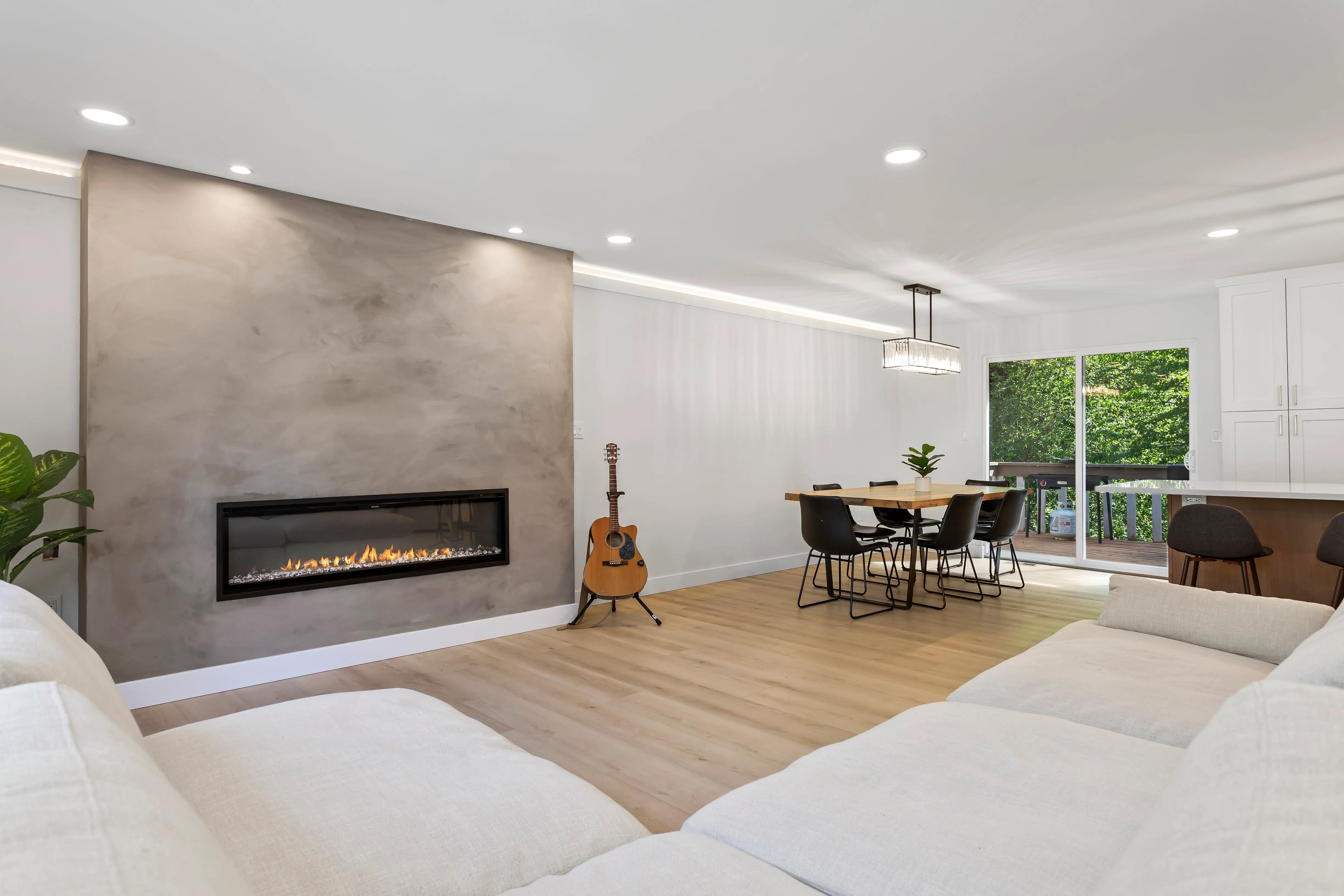 Open living and dining room with light wood flooring, modern linear fireplace, acoustic guitar, black dining chairs, and sliding glass door with green outdoor view.