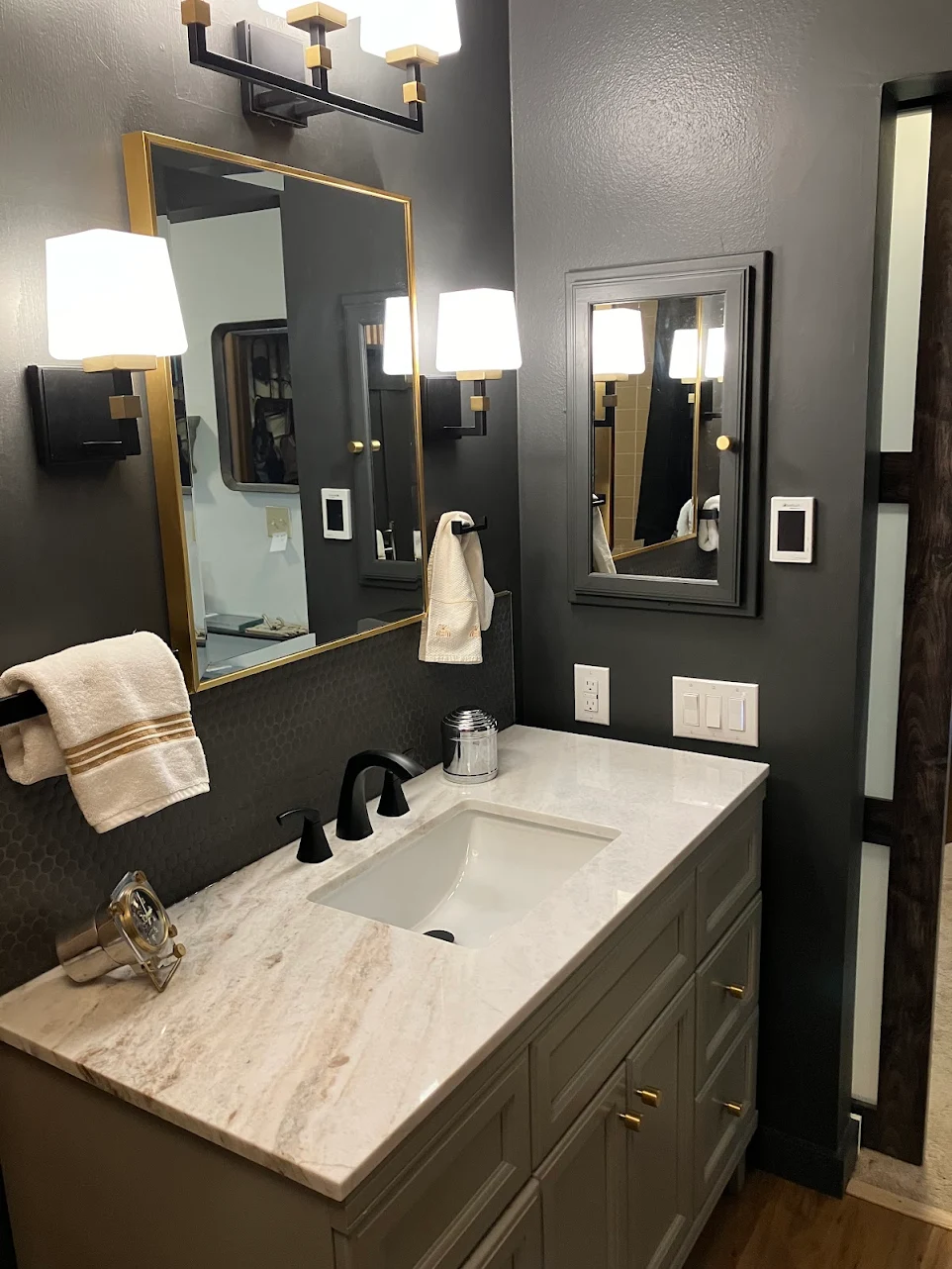 Modern bathroom vanity with marble countertop, black faucets, gold-framed mirror, and wall-mounted light fixtures.