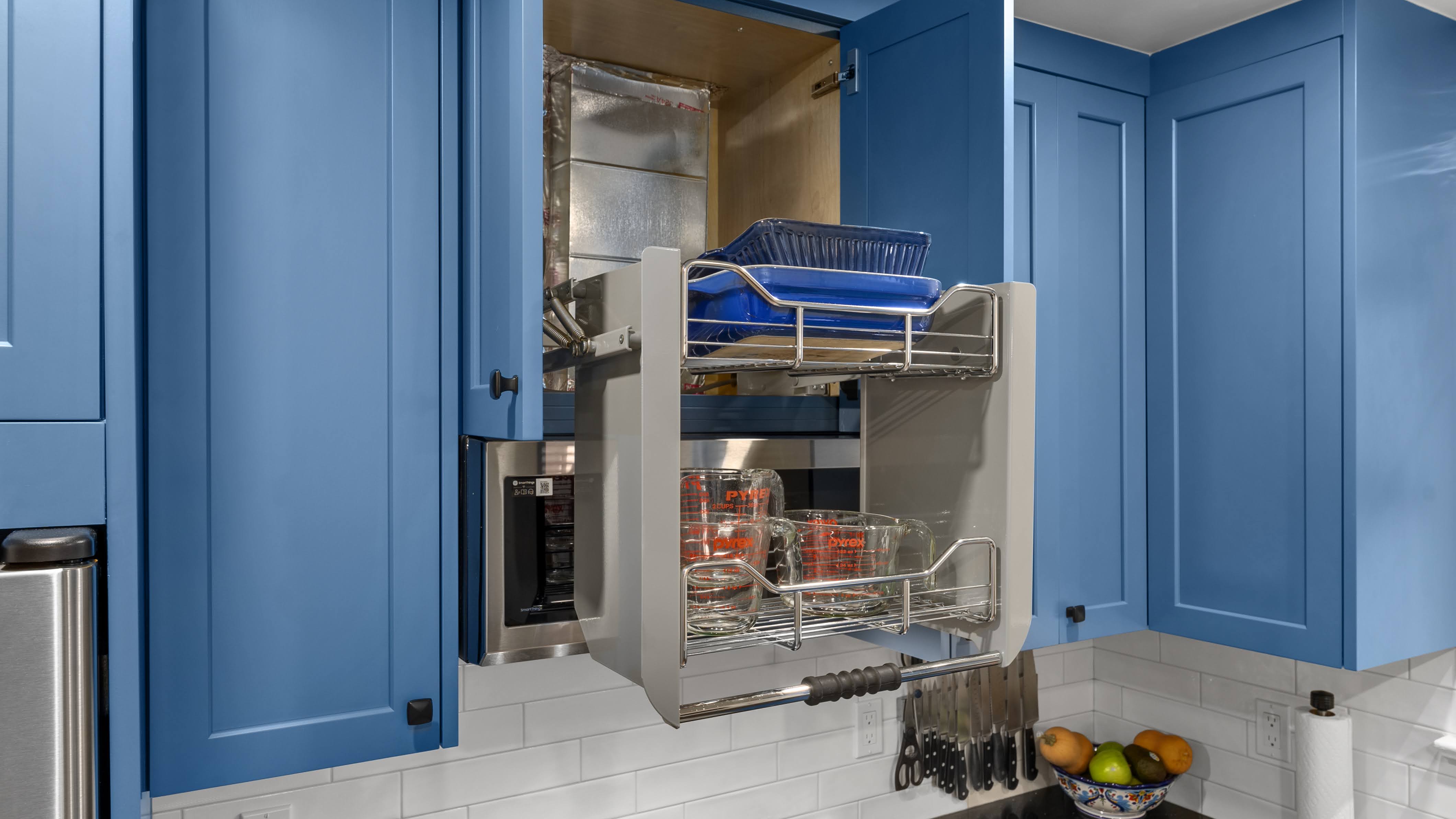 Pull-down kitchen cabinet organizer inside blue cabinets holding glass measuring cups and blue trays.