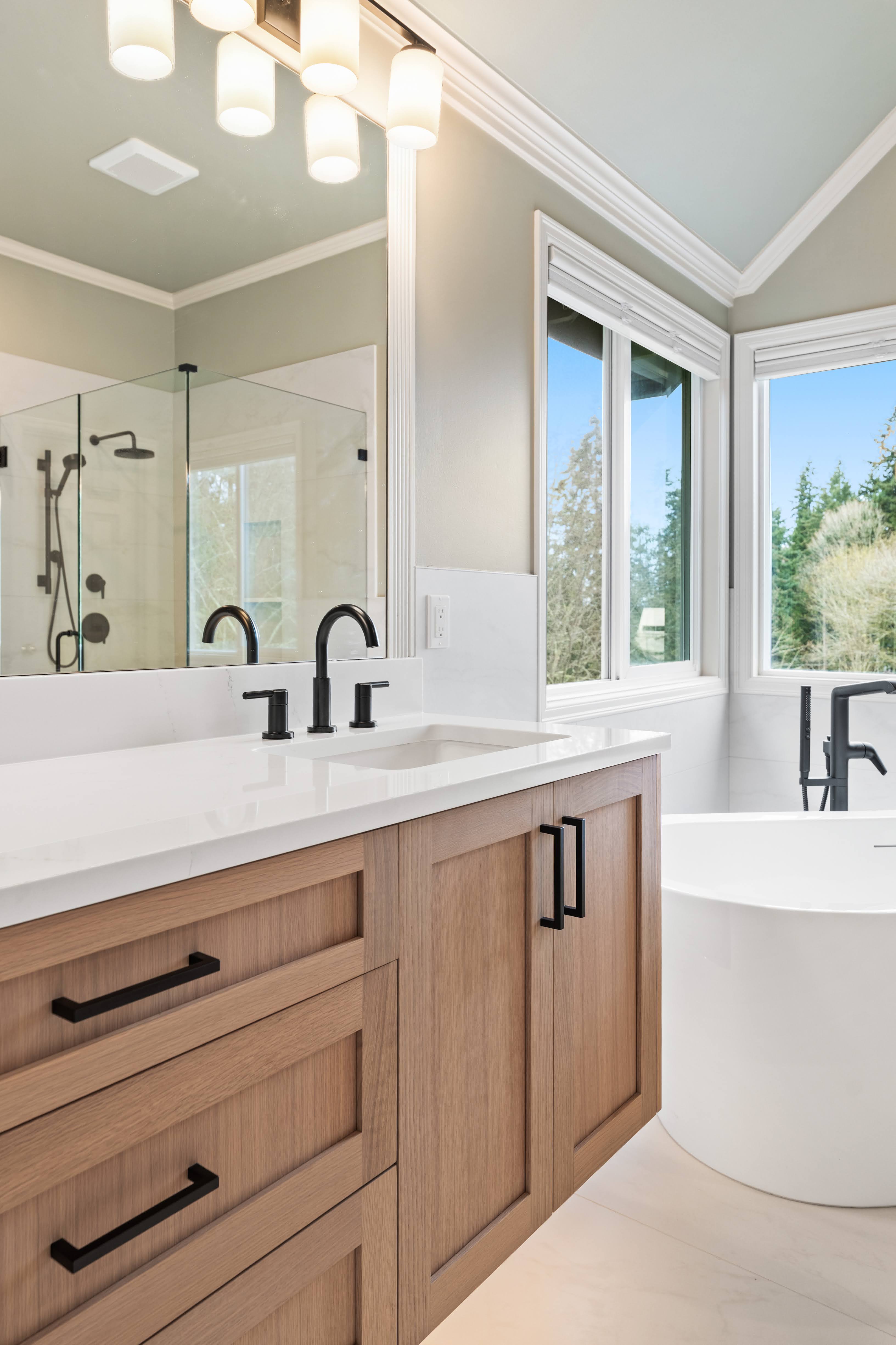 Light wood bathroom vanity with black fixtures next to a sleek soaking tub and large windows with outdoor views