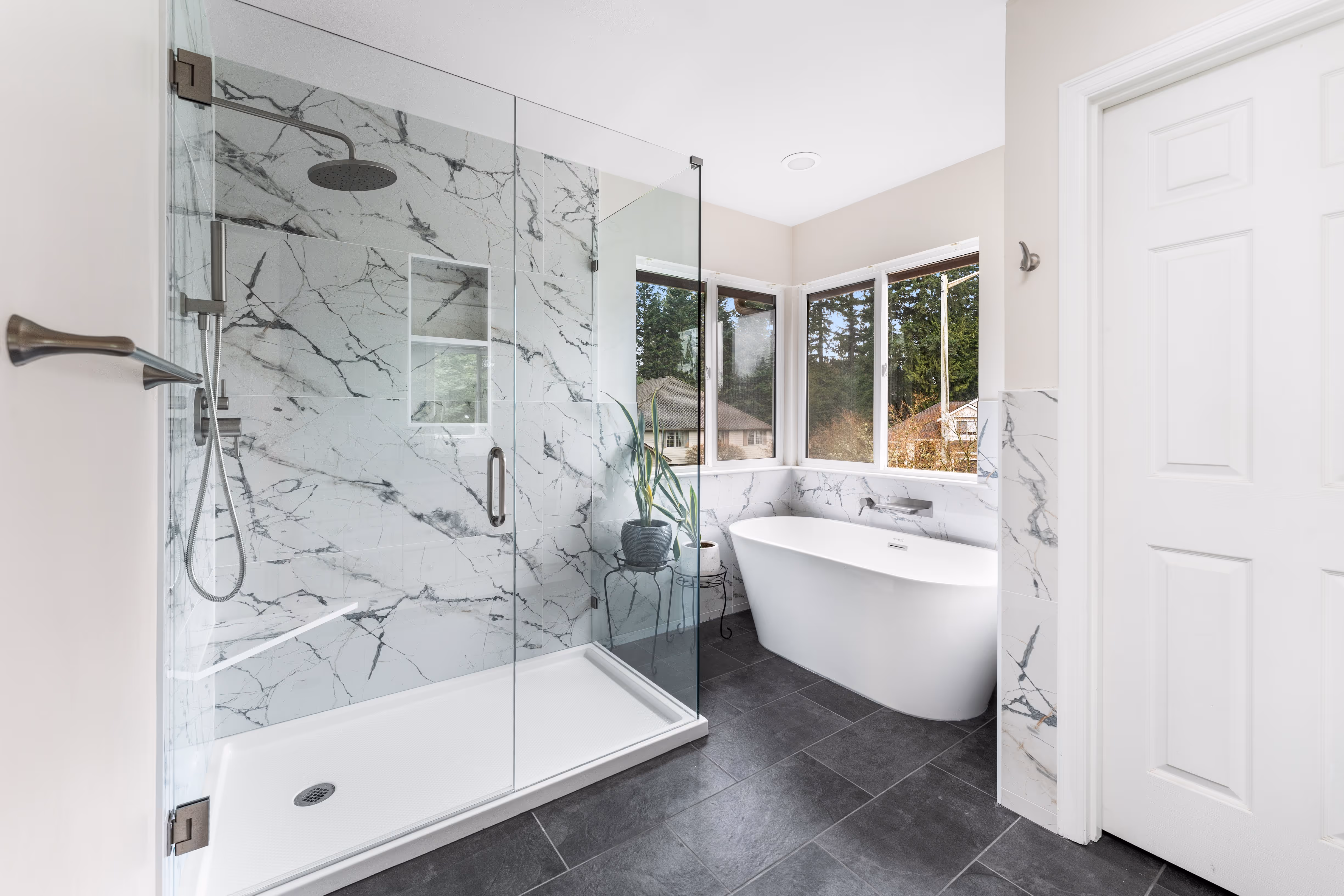 Bright modern bathroom with marble-tiled walk-in shower, freestanding bathtub by corner windows, and dark floor tiles.