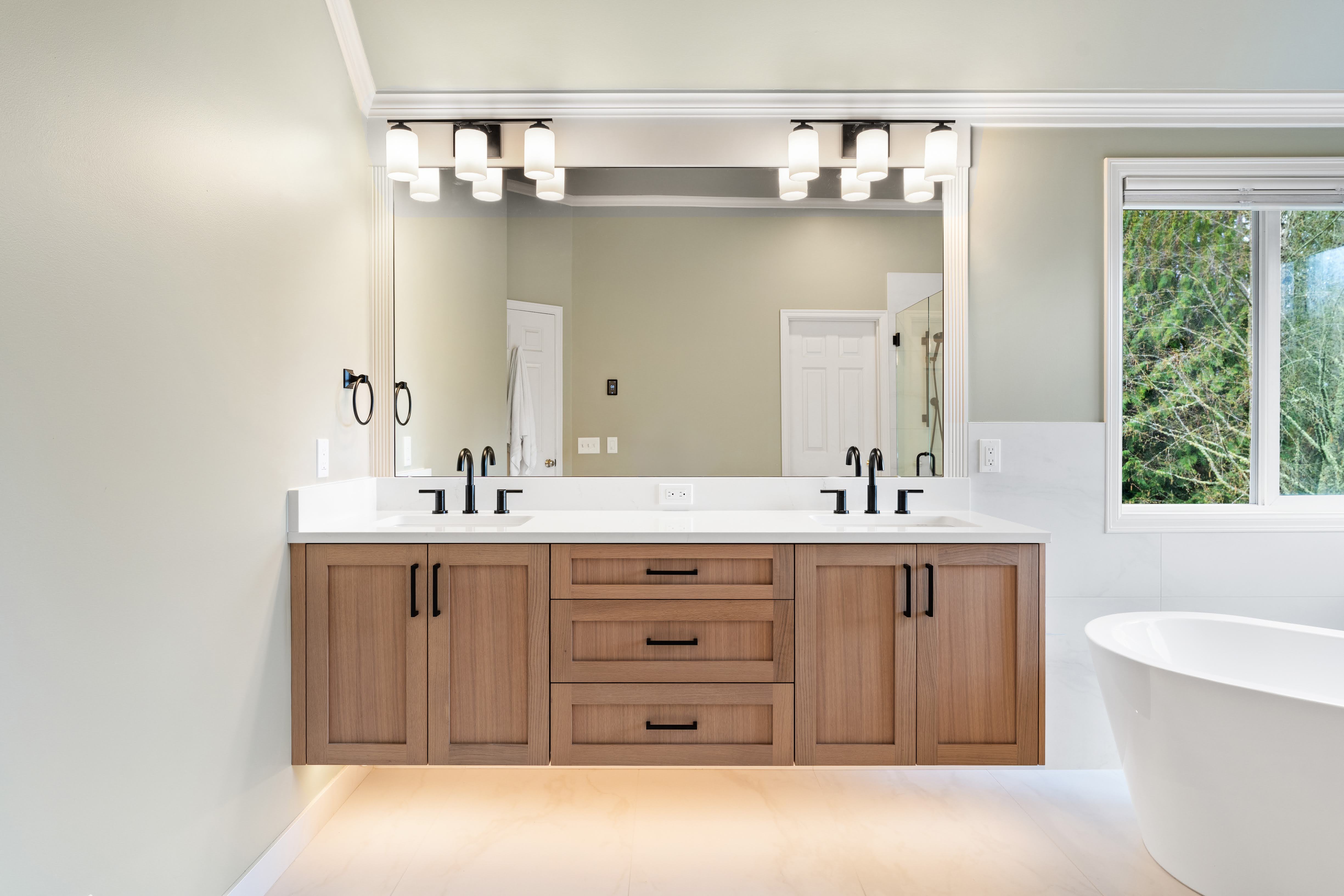 bathroom with a floating wood double vanity, black faucets, large framed mirror, soft overhead lighting, freestanding bathtub, and a window overlooking greenery