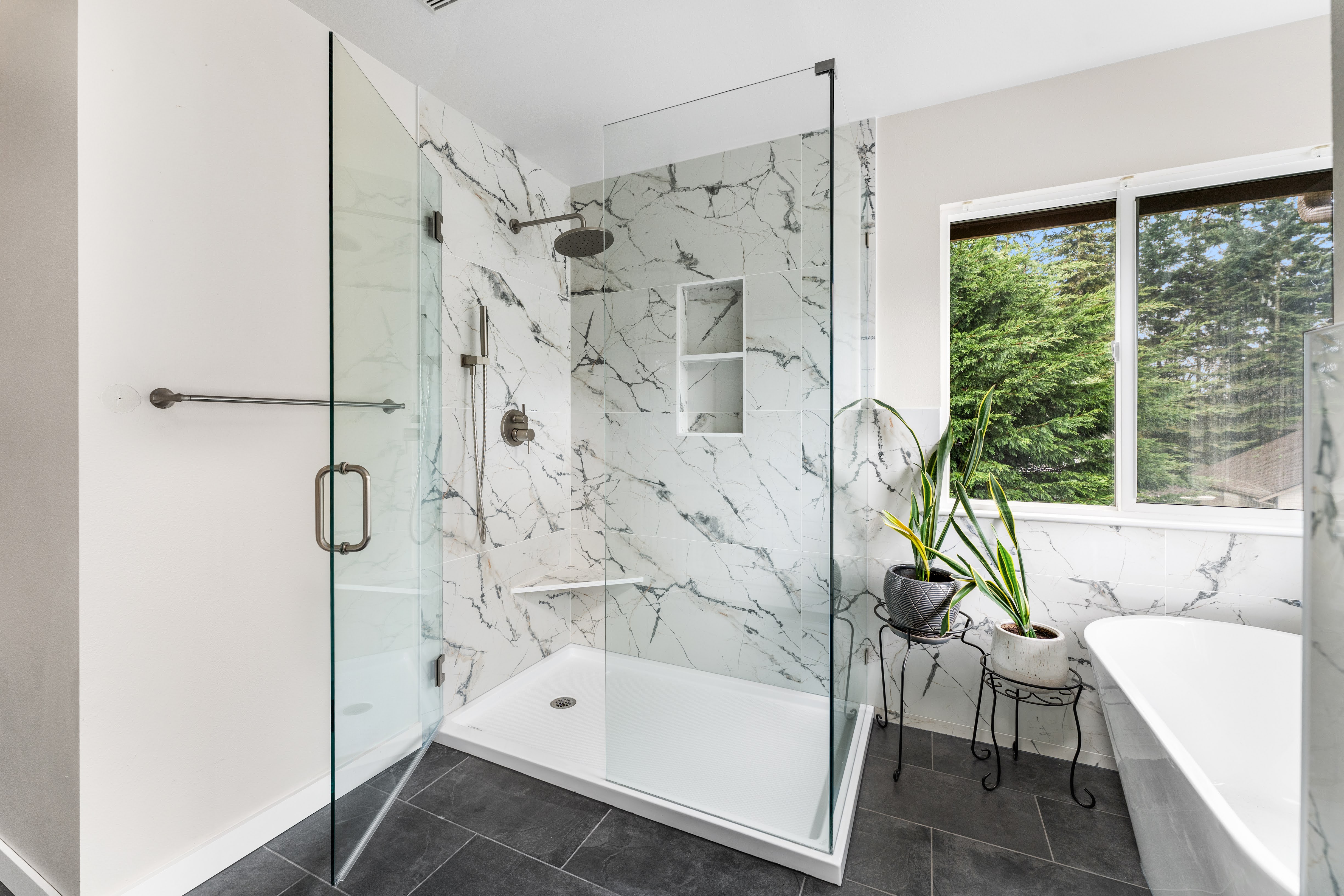 Contemporary bathroom featuring a frameless glass shower, large marble-look tiles, built-in shower shelves, dark floor tiles, and a freestanding tub near a bright window