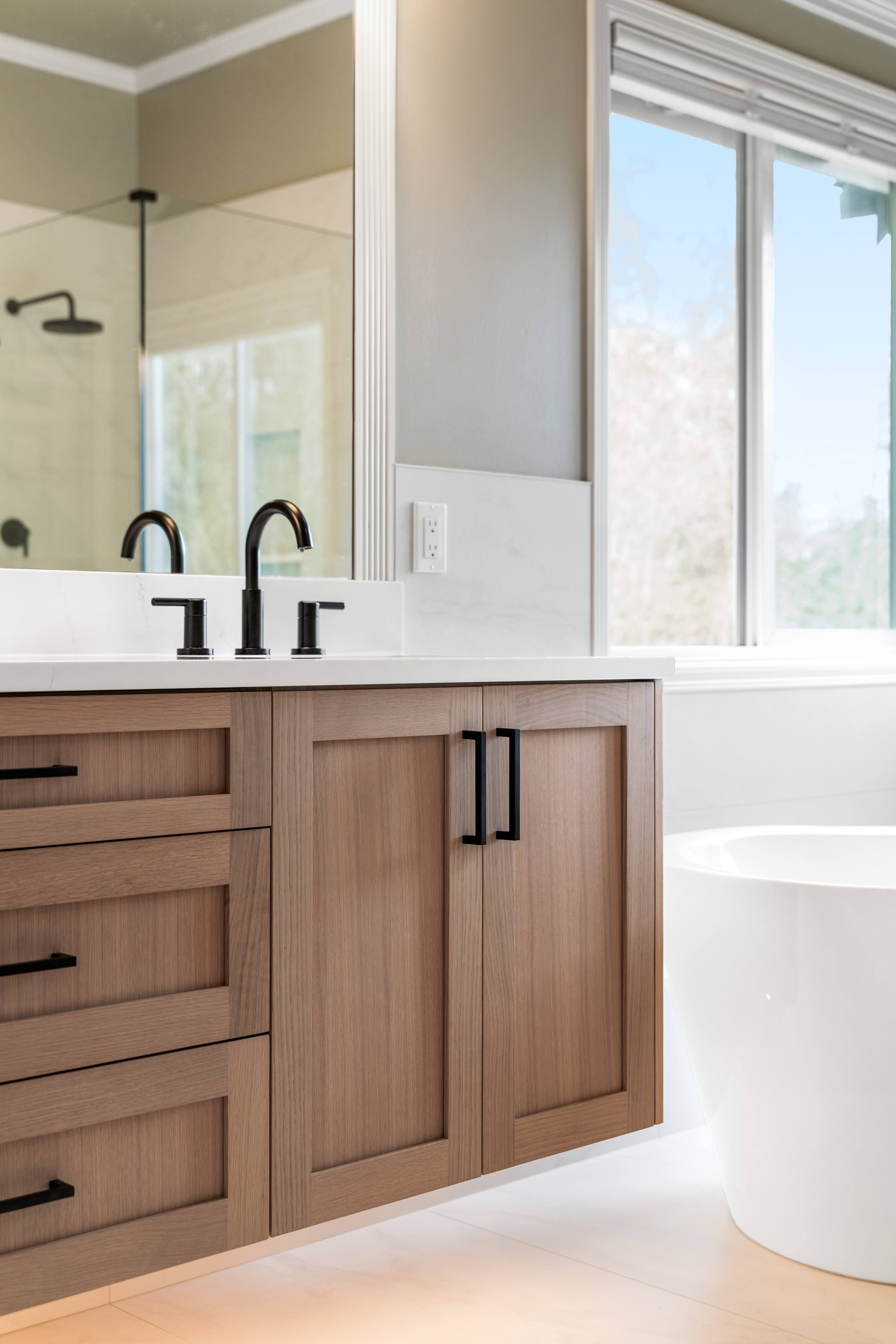 Modern bathroom vanity with light wood cabinets, black faucet, and a large mirror reflecting a shower area, next to a white freestanding bathtub by a window.