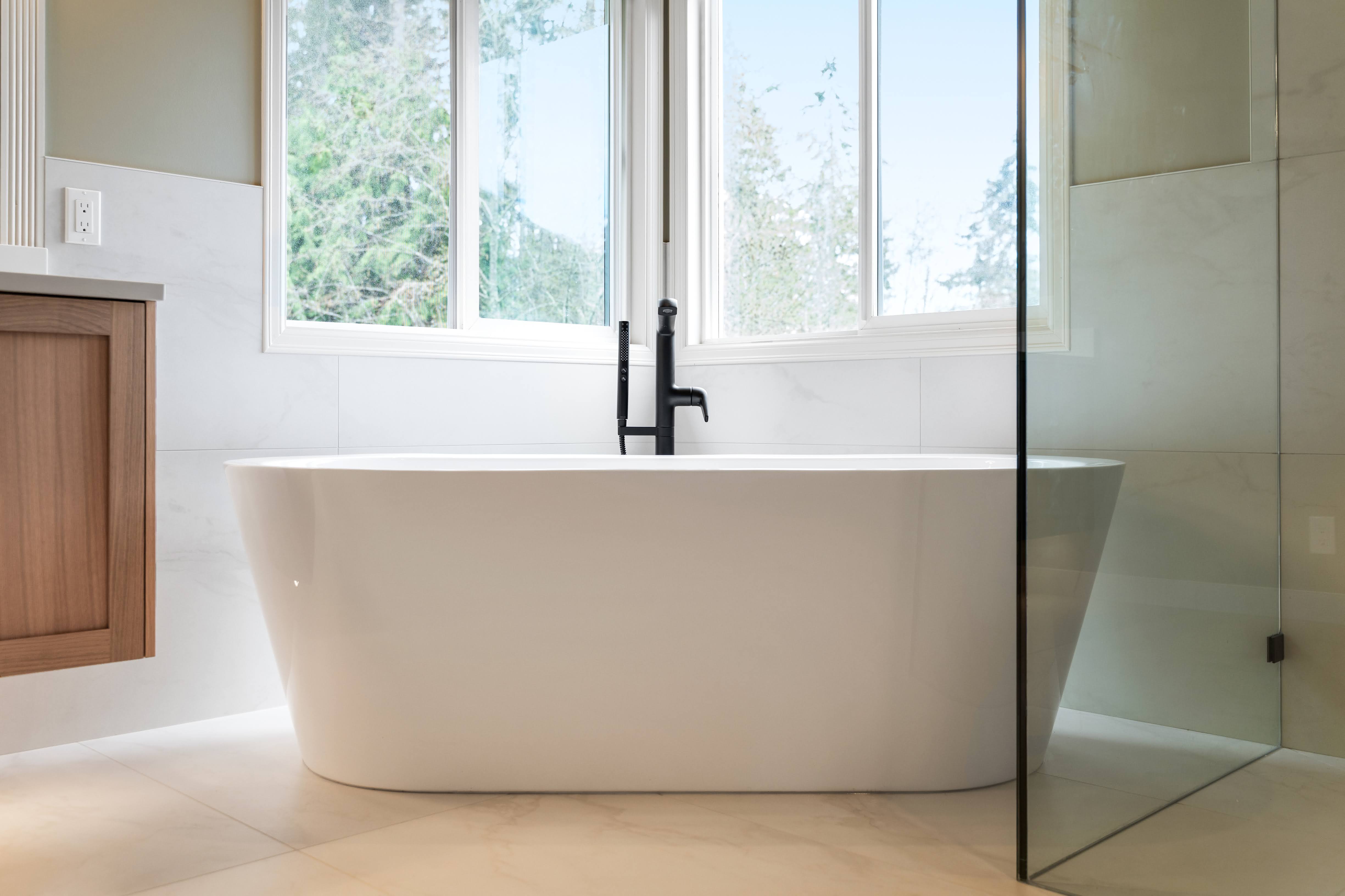 Modern white freestanding bathtub under large windows with a black faucet in a bright bathroom.