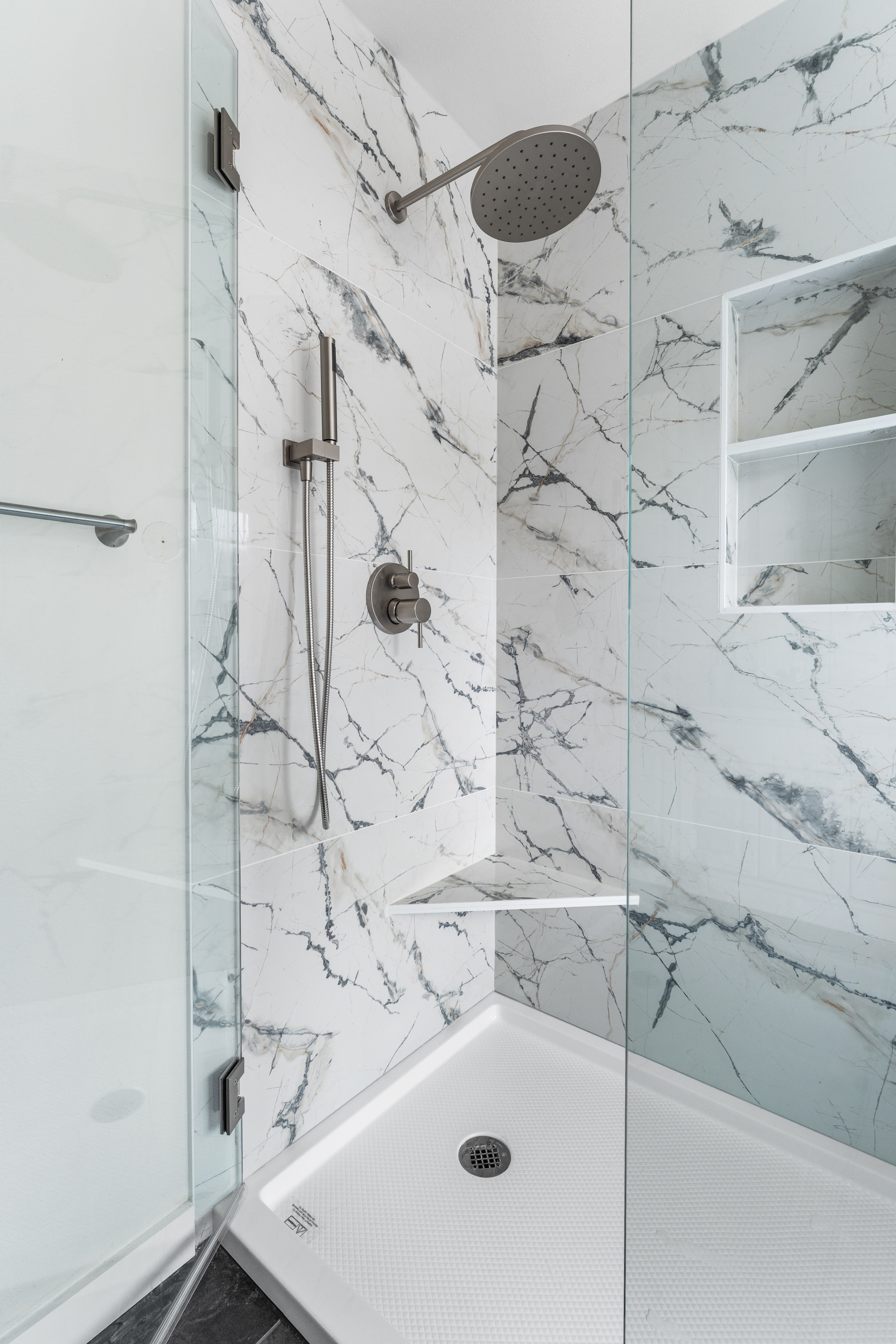 Modern shower with marble walls, a rain showerhead, handheld shower, corner shelf, and glass door.