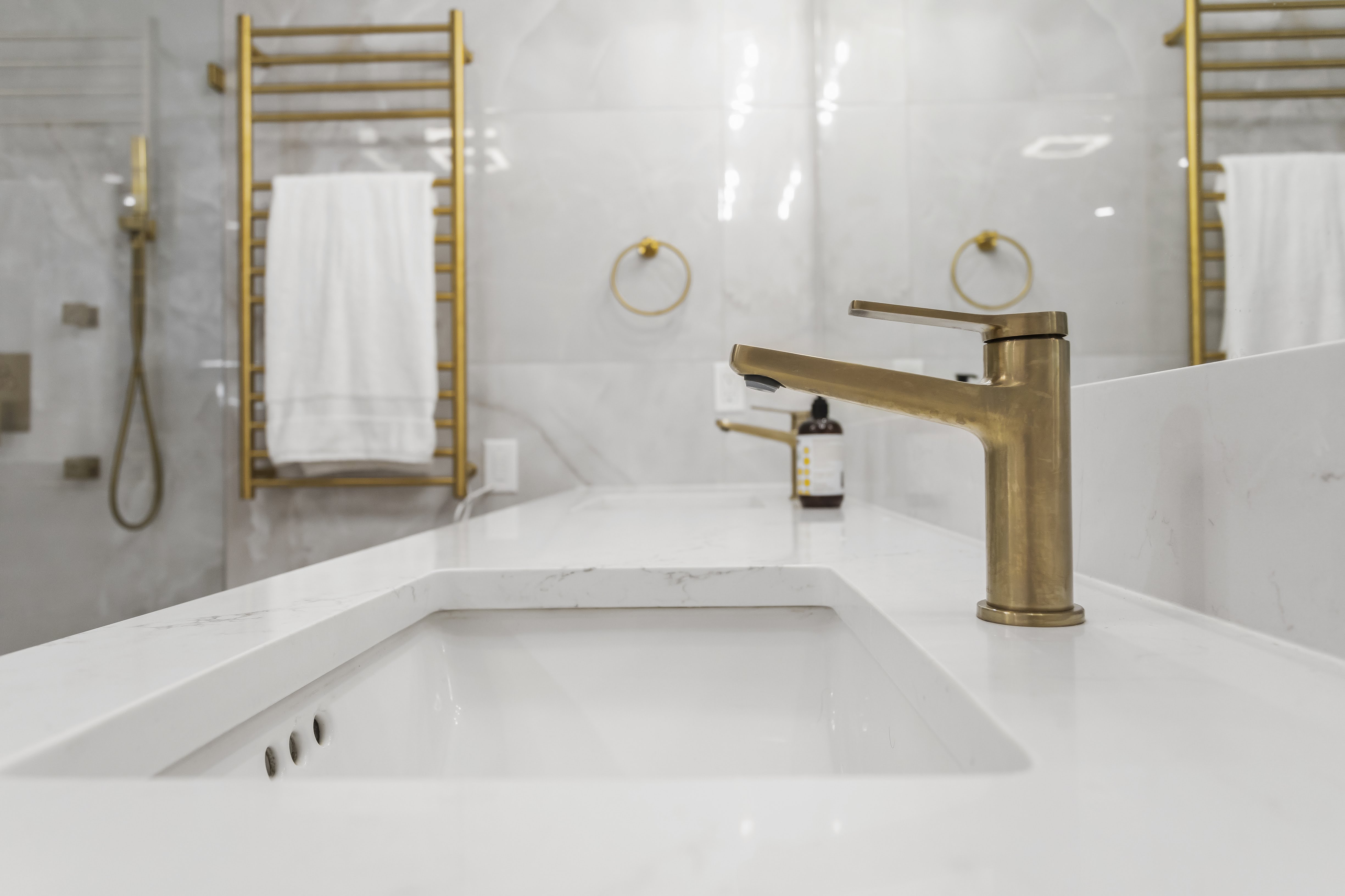 Modern bathroom sink with white marble countertop and gold faucet, towel rack, and mirror in the background.