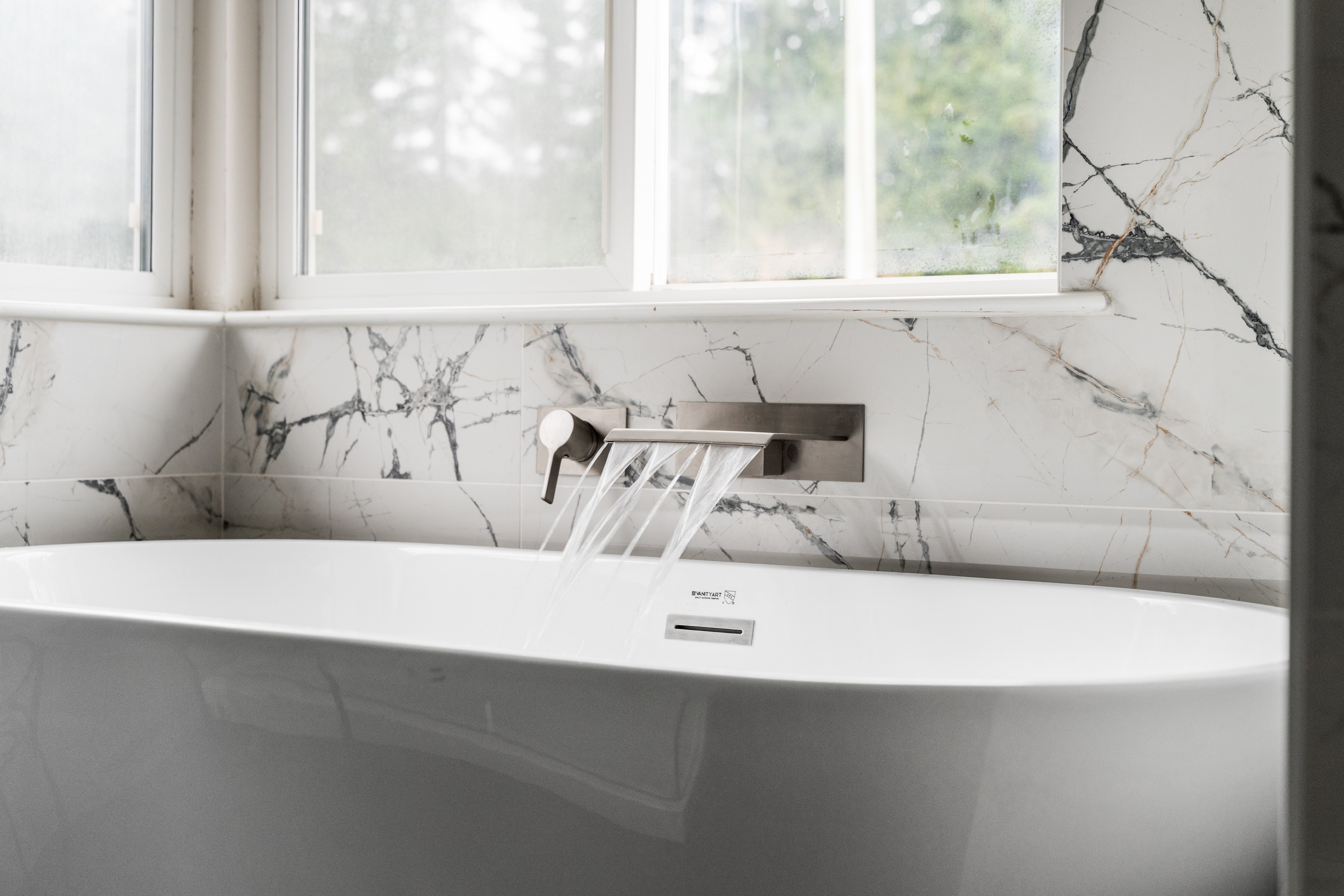 Modern bathroom bathtub with a wide spout faucet flowing water, set against white marble walls and a frosted window.