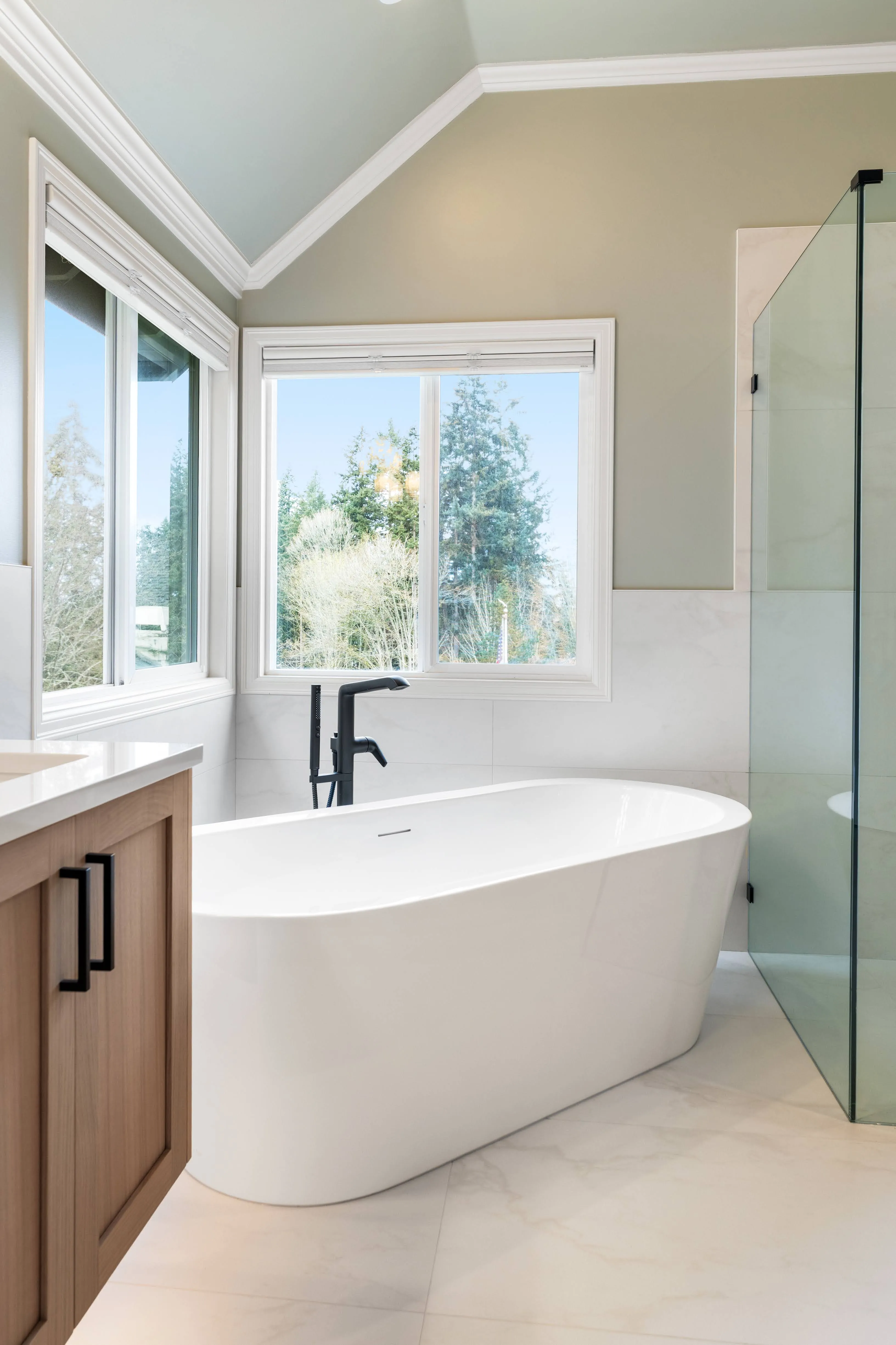 Freestanding bathtub positioned under large windows in a modern bathroom with light tile floors, black fixtures, and glass shower panels
