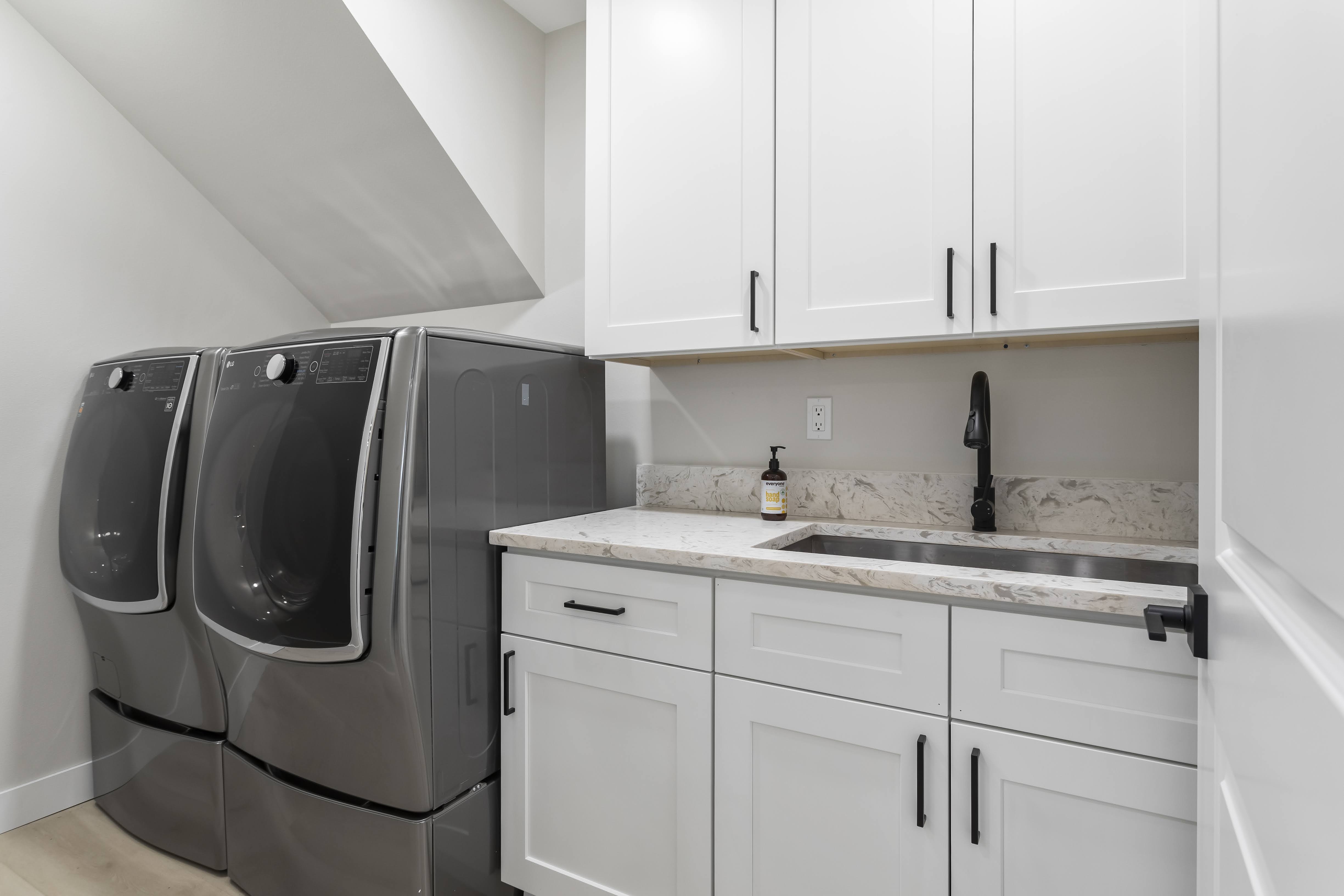 Contemporary laundry room with grey front-loading washer and dryer, white cabinetry with black handles, marble countertop, and black faucet over a stainless steel sink.