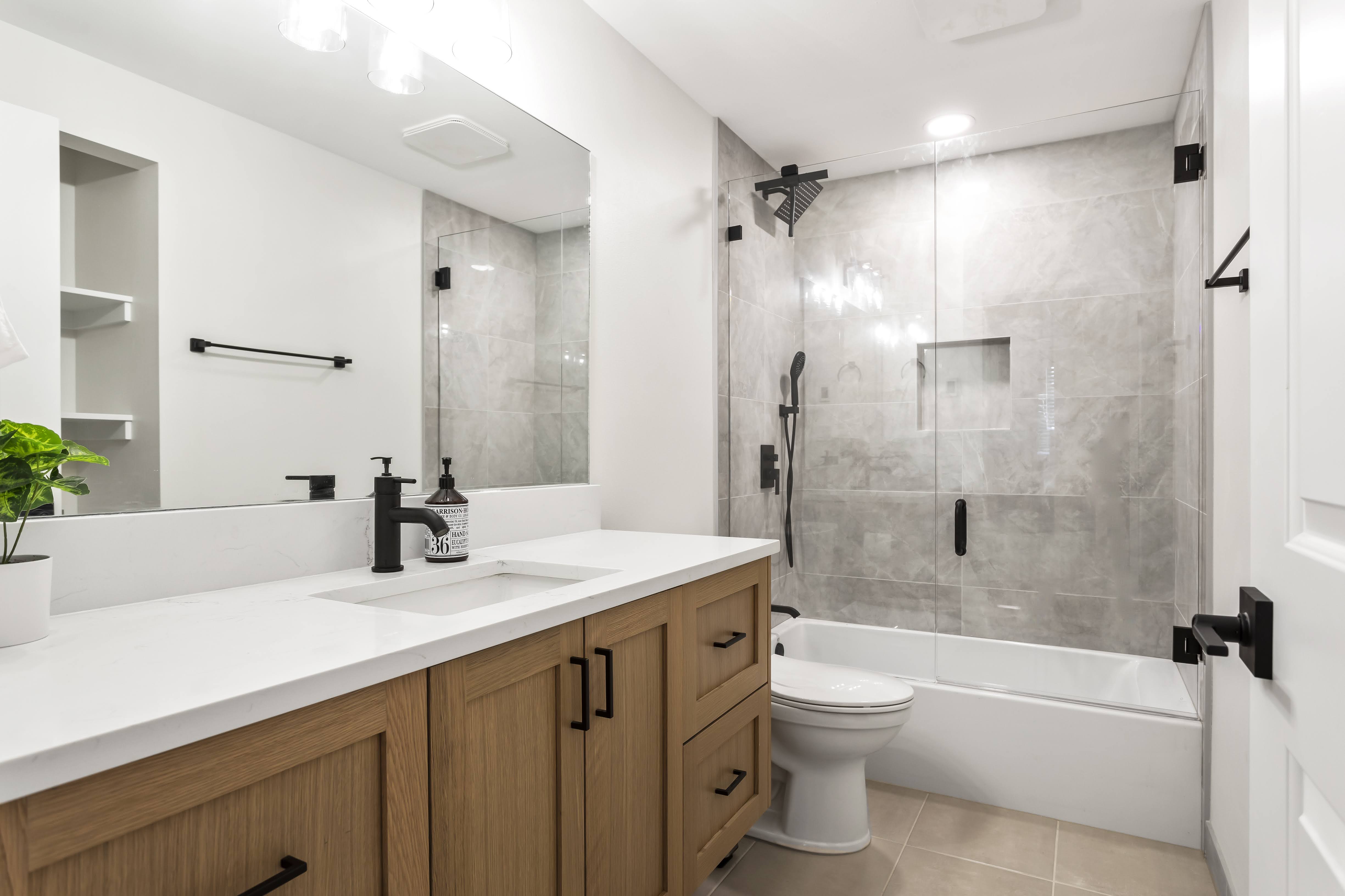 Modern bathroom with wooden vanity, white countertop, black fixtures, a large mirror, and a glass-enclosed bathtub with gray tiled walls.