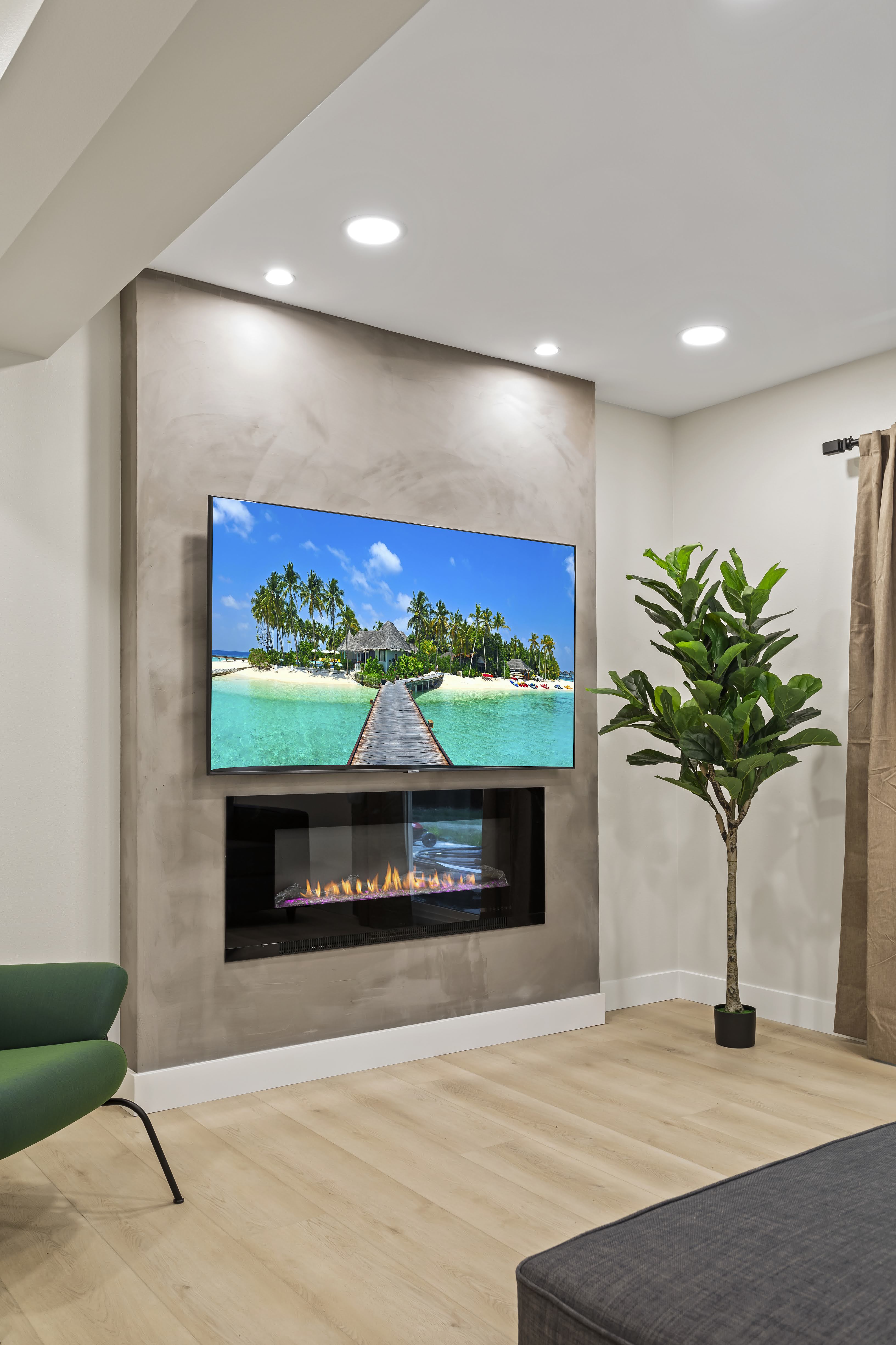Modern living room corner with a wall-mounted TV displaying a tropical island, electric fireplace below, large green plant, and hardwood floor.