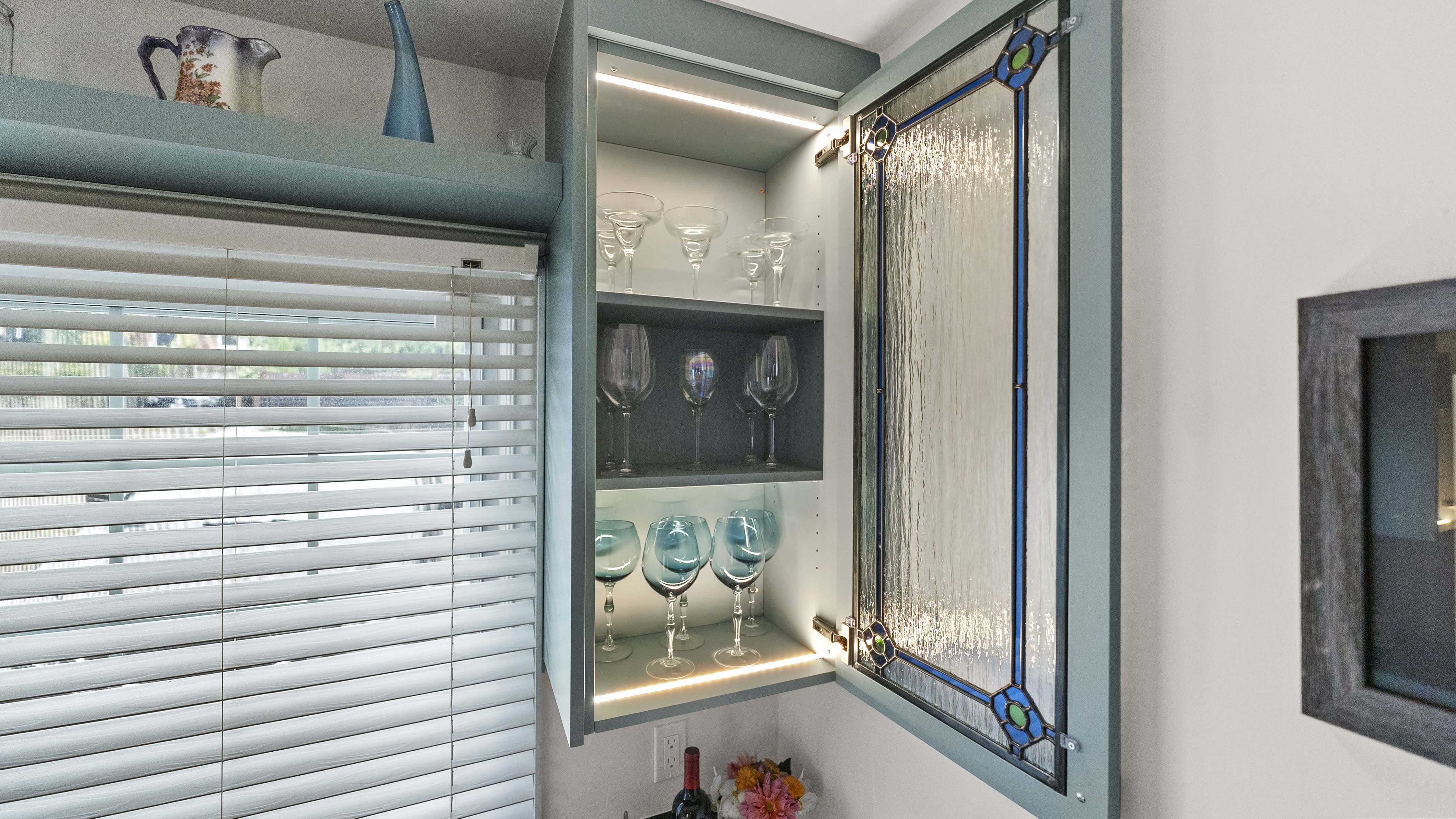 Open teal kitchen cabinet with stained glass door showing shelves with various wine glasses and cocktail glasses inside, next to a window with white blinds.
