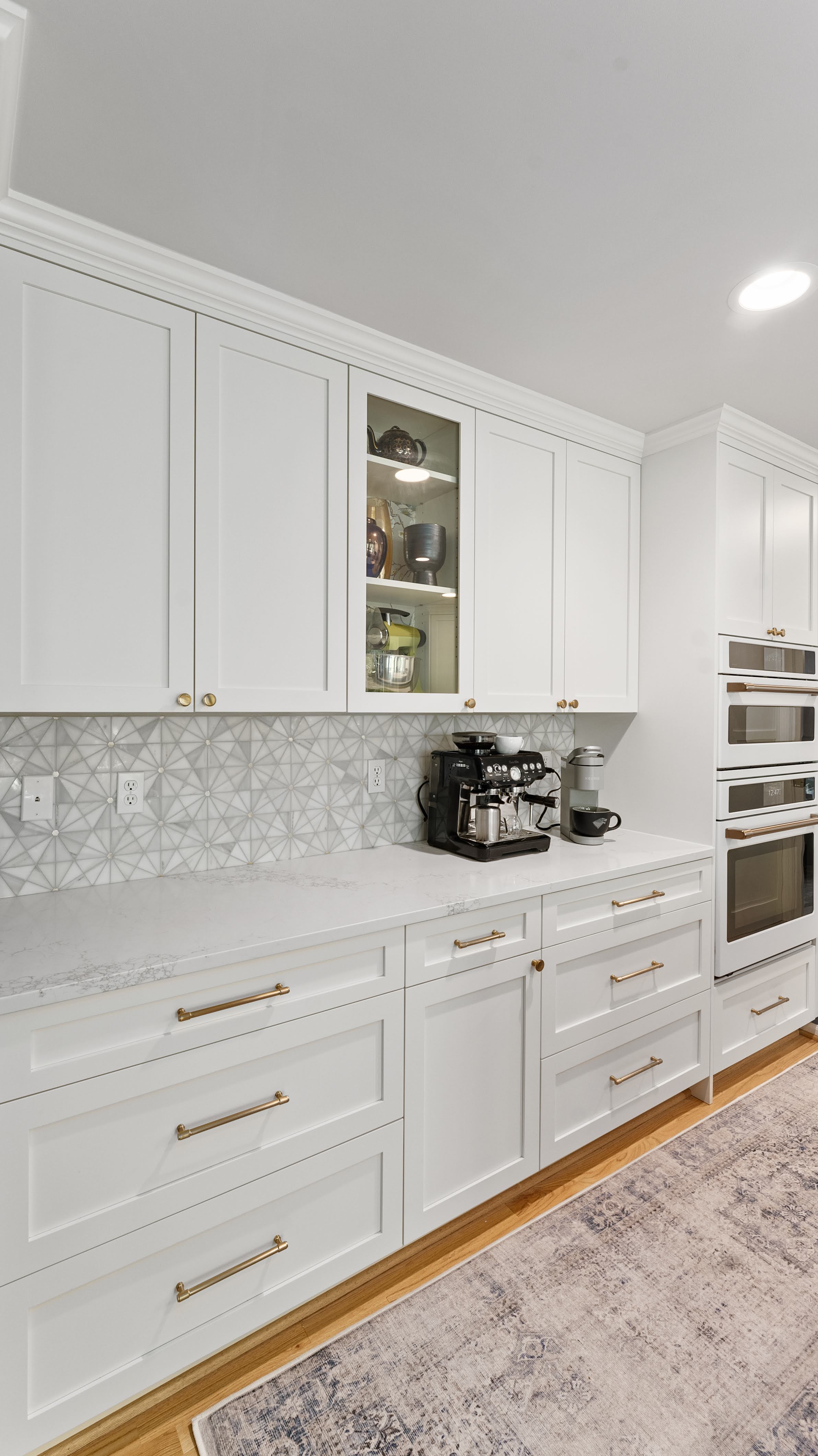 Bright modern kitchen with white shaker cabinets, gold hardware, patterned tile backsplash, quartz countertops, built-in double ovens, and a coffee station setup