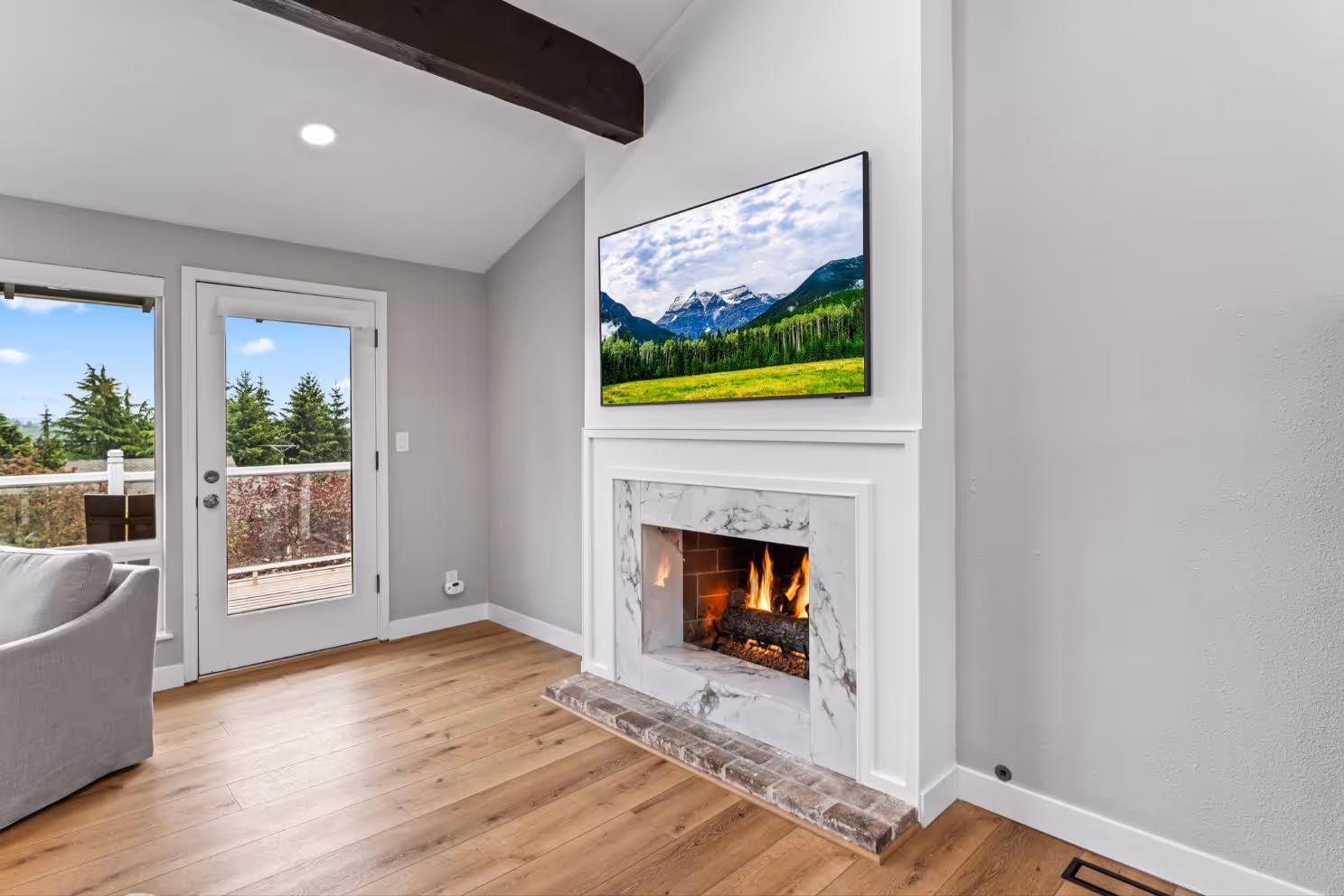 Cozy living room with a marble fireplace, mounted TV, light wood flooring, and glass doors leading to an outdoor deck with scenic tree views