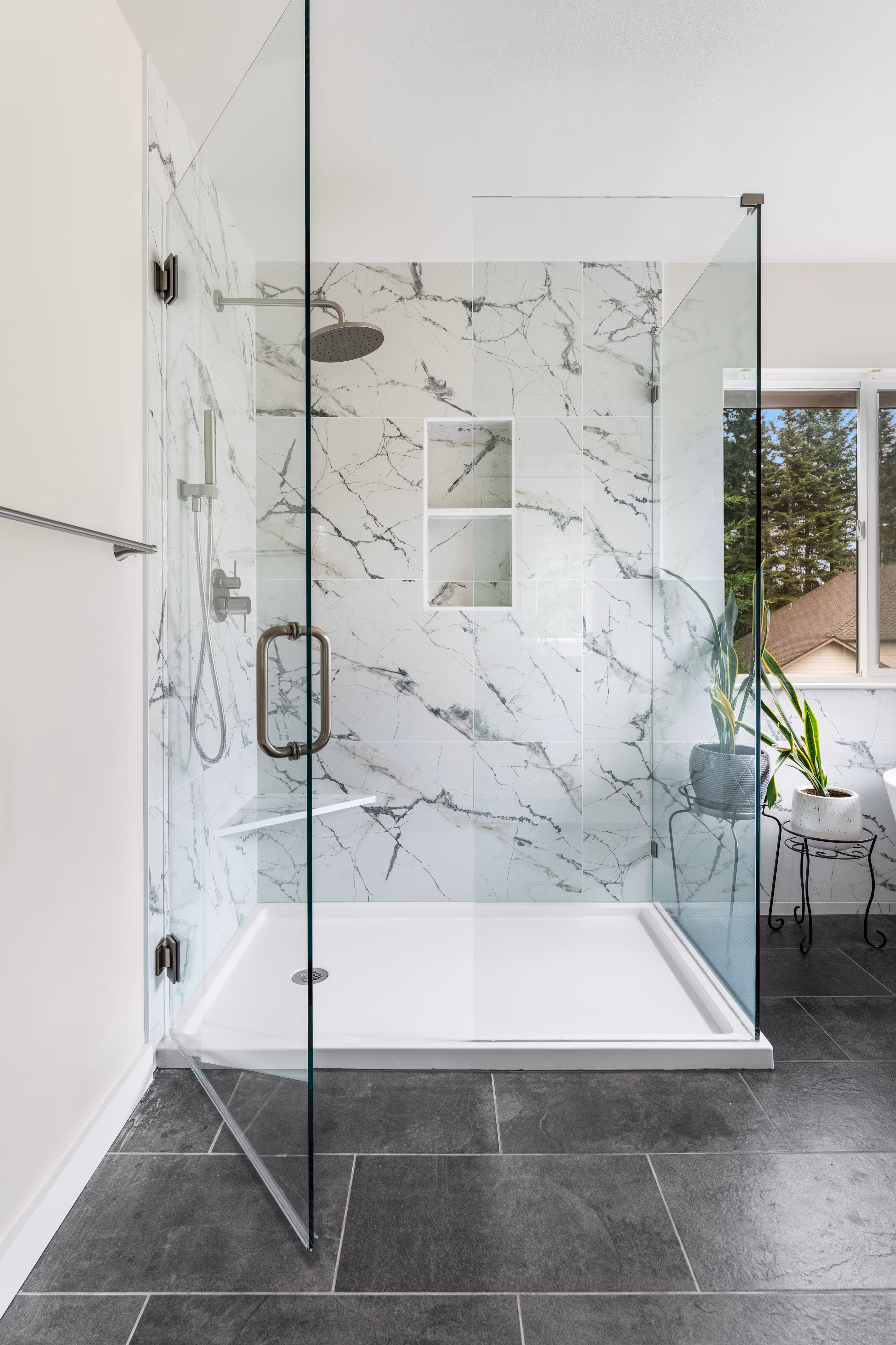 bathroom with a glass walk-in shower featuring white marble-pattern tile, built-in niches, matte black fixtures, and dark slate flooring beside a window with natural light and potted plants