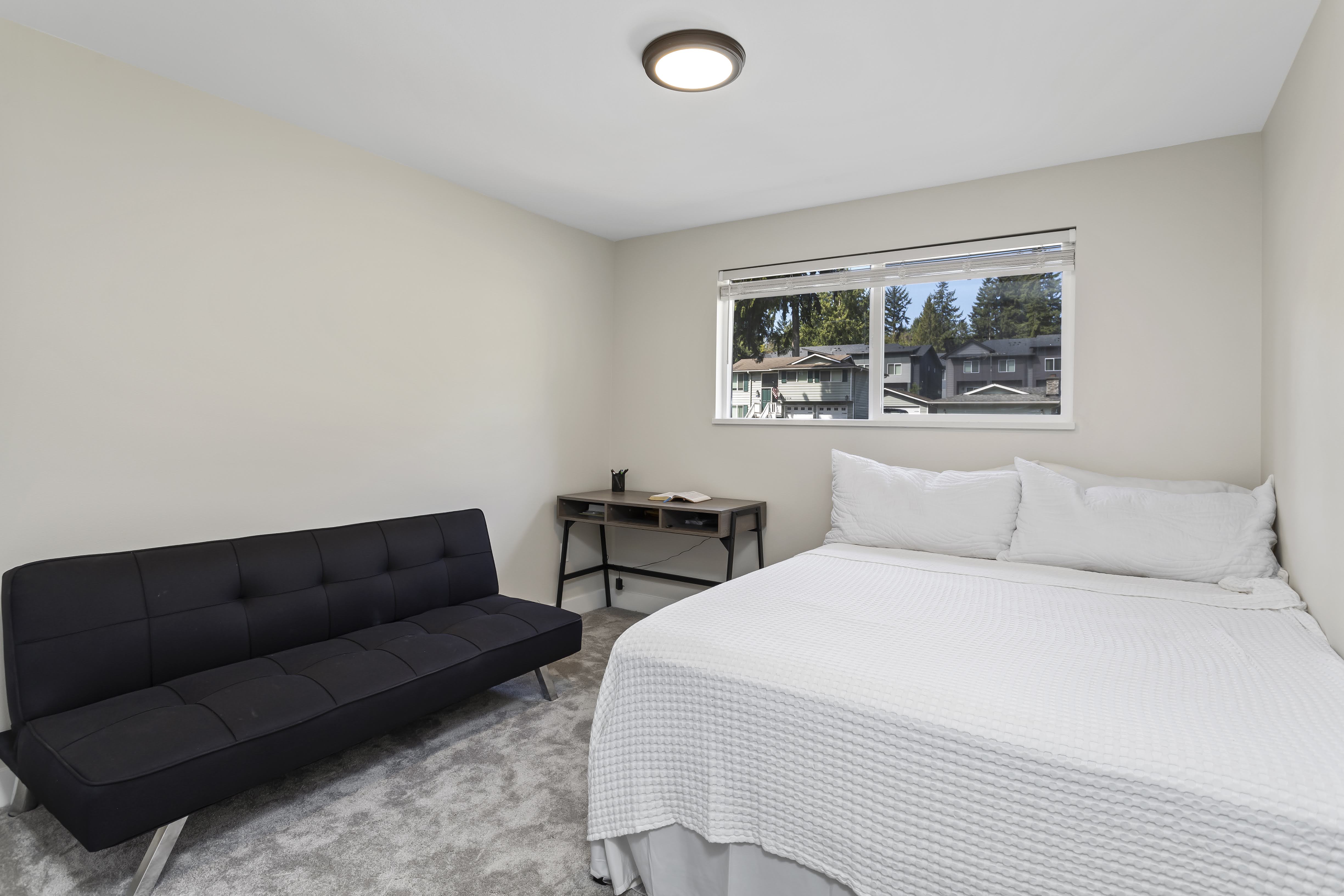 Simple modern bedroom with a white bed, black futon, small desk, neutral walls, and a large window letting in natural light with views of nearby homes and trees