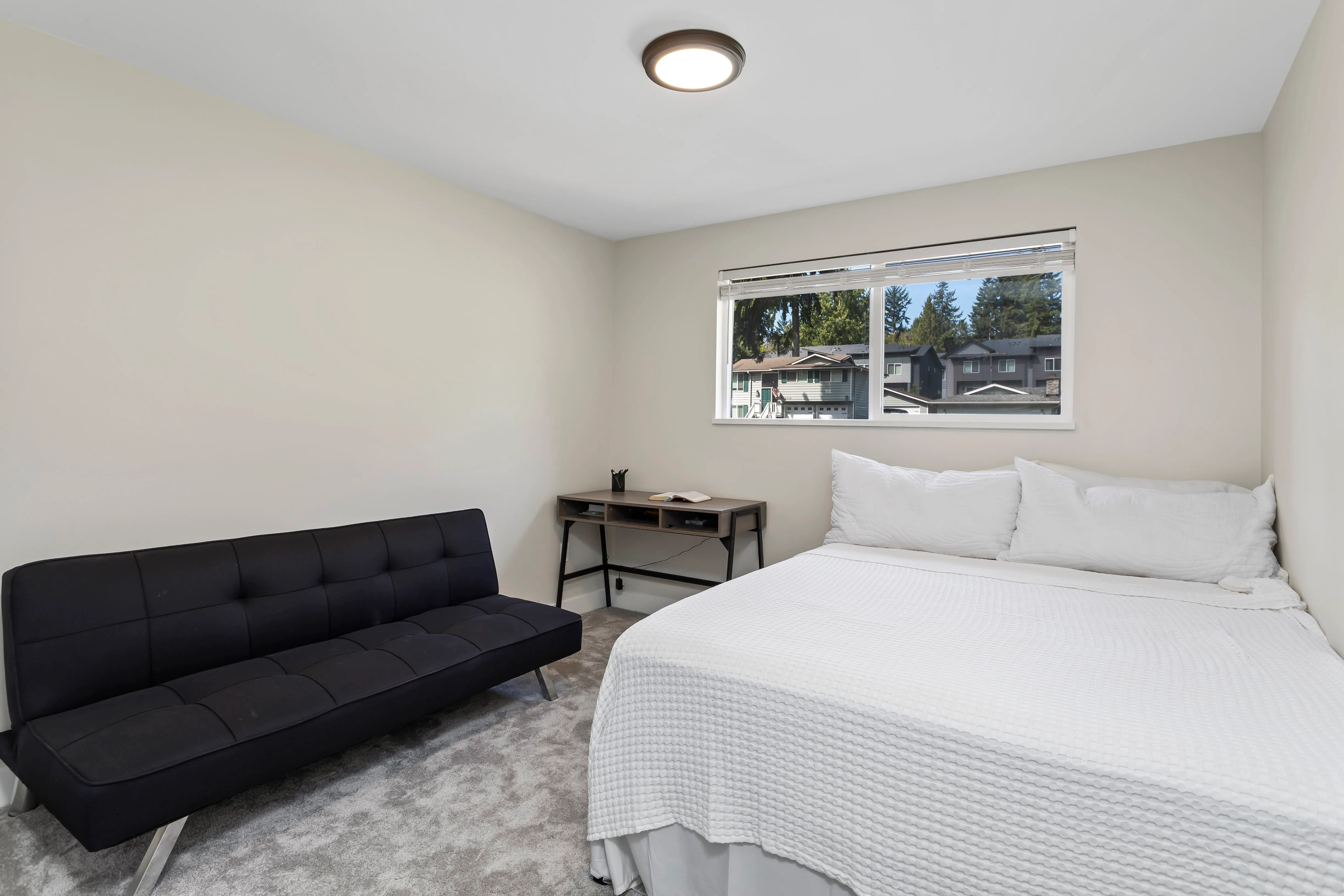 Simple modern bedroom with a white bed, black futon, small desk, neutral walls, and a large window letting in natural light with views of nearby homes and trees