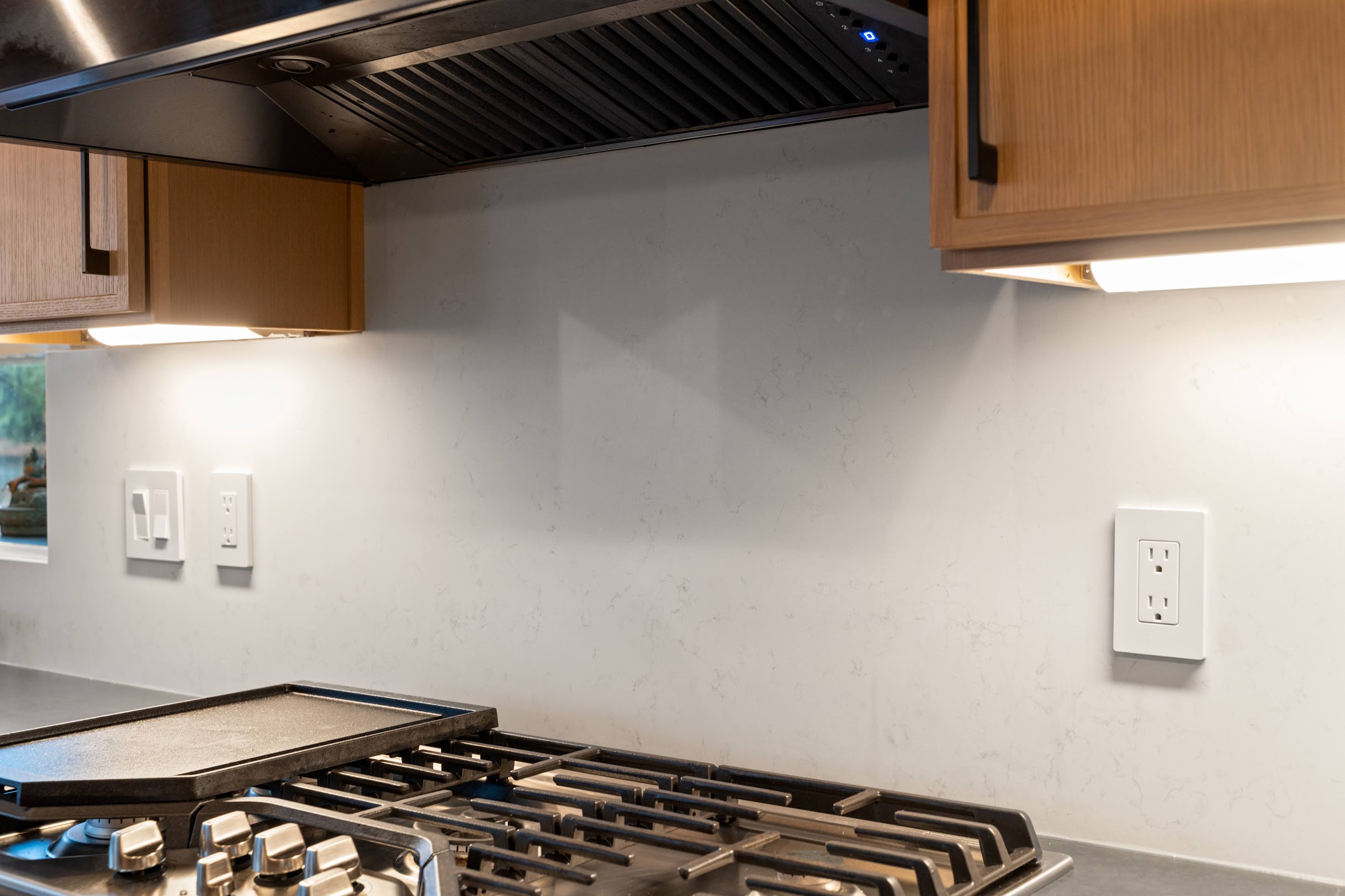 White quartz slab backsplash with subtle veining behind gas stovetop, natural wood cabinets, and black range hood in modern kitchen remodel by Crafthaus