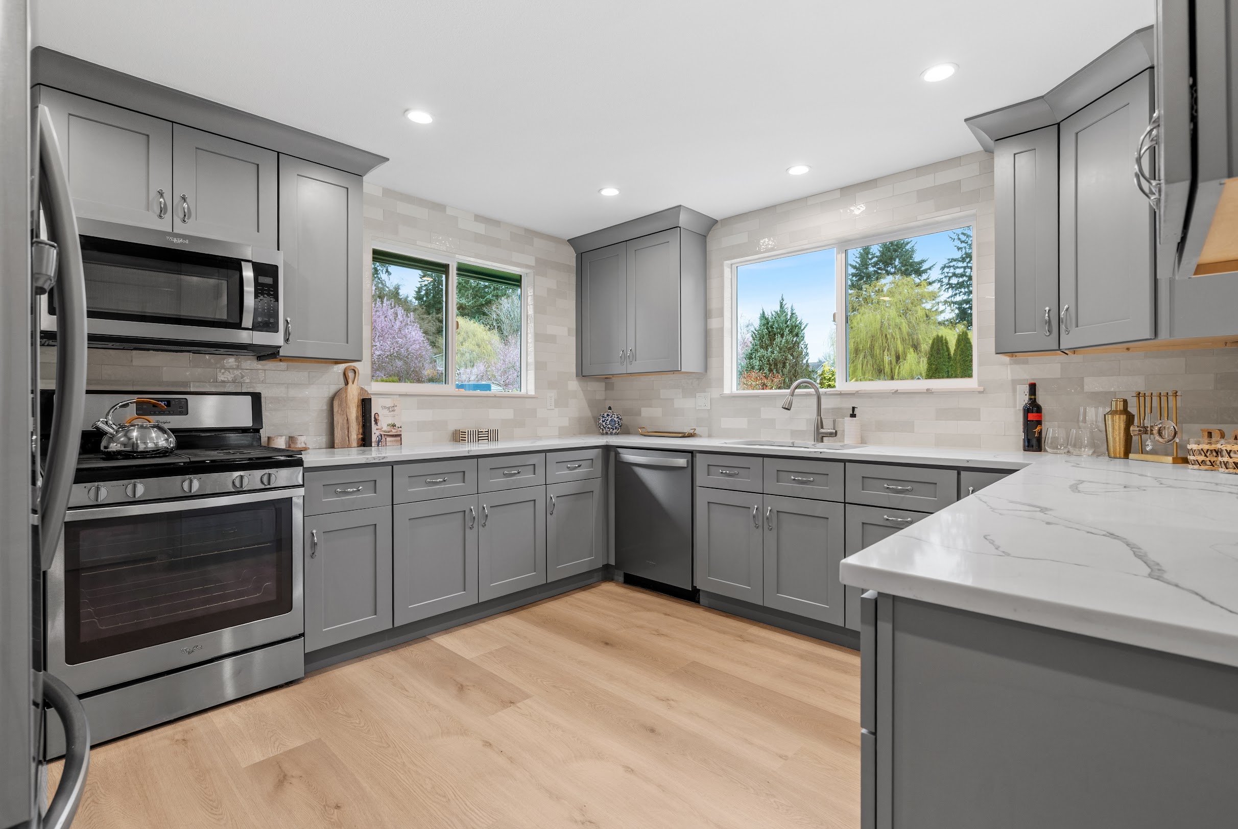 Wide-angle view of full gray shaker cabinet kitchen remodel with cream rectangular tile backsplash, white quartz countertops, stainless appliances, and garden windows by Crafthaus