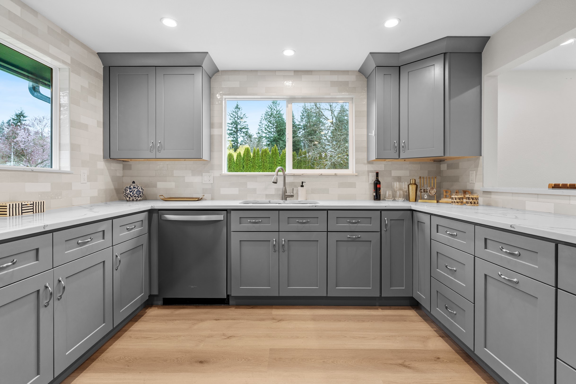 Full kitchen view of cream brick-style tile backsplash with gray shaker cabinets, white quartz countertops, double sink, and hardwood floors in U-shaped layout by Crafthaus