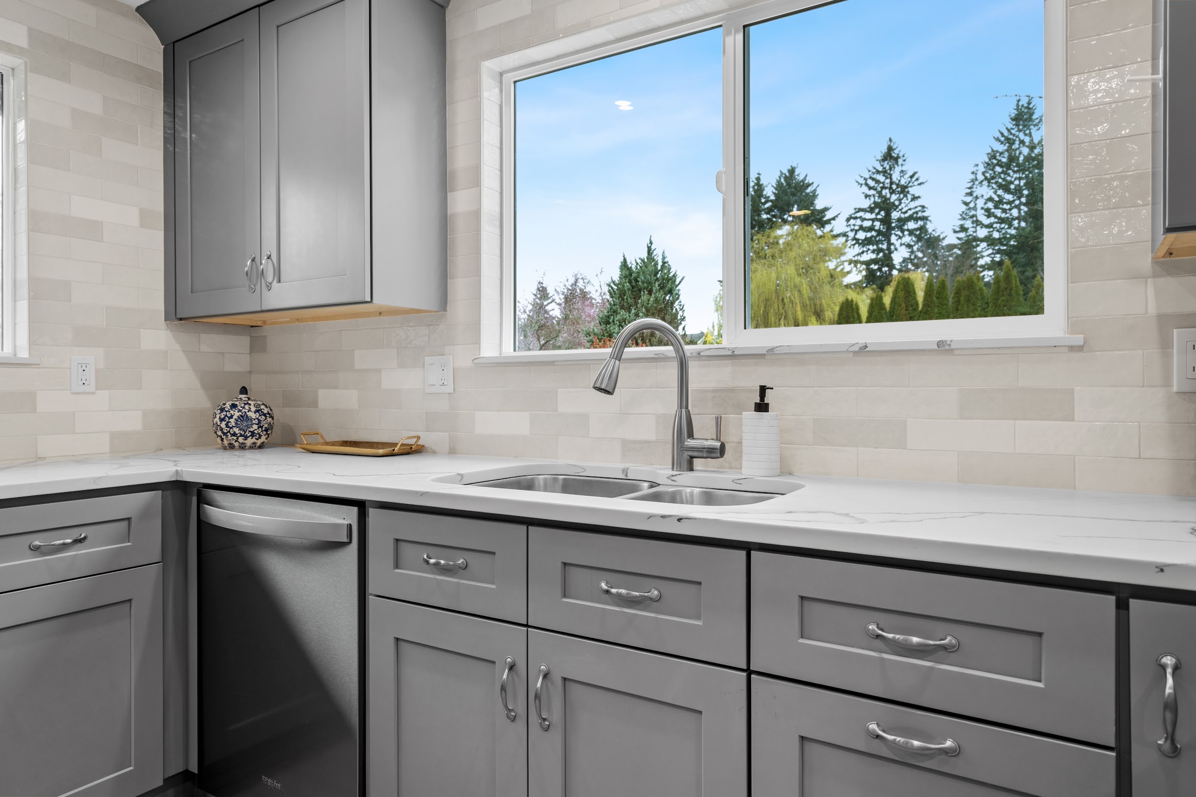 Cream and beige rectangular tile backsplash with gray shaker cabinets, white quartz countertop, and double undermount sink with garden window view, Crafthaus kitchen remodel
