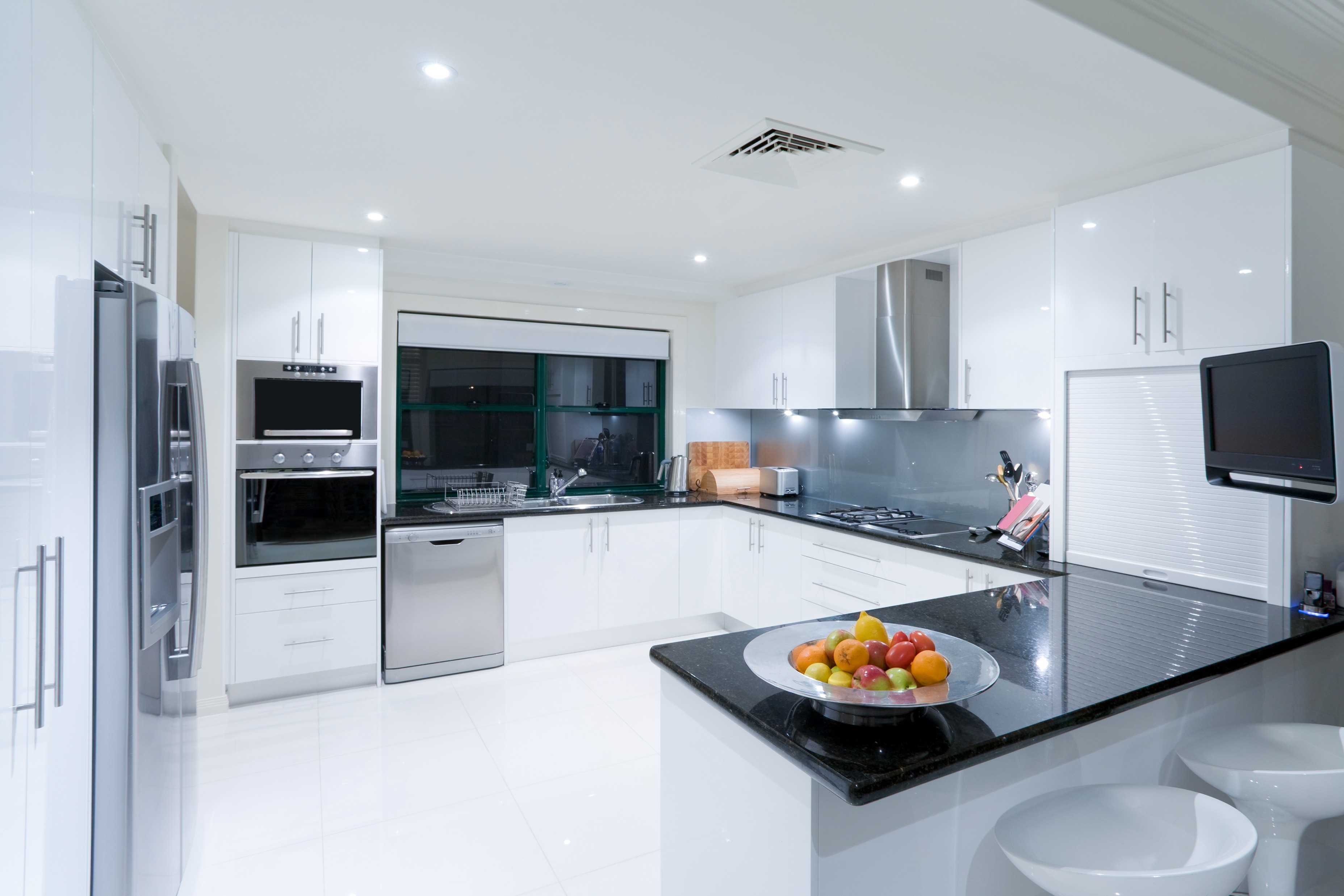 Modern white kitchen with glossy cabinets, black granite countertops, stainless steel appliances, a built-in oven and refrigerator
