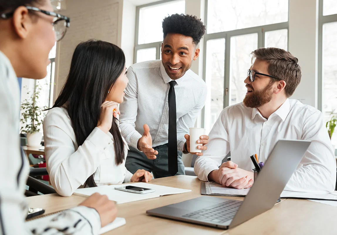 Four diverse colleagues in a bright office engaged in a friendly discussion about outsourced advertising sales.