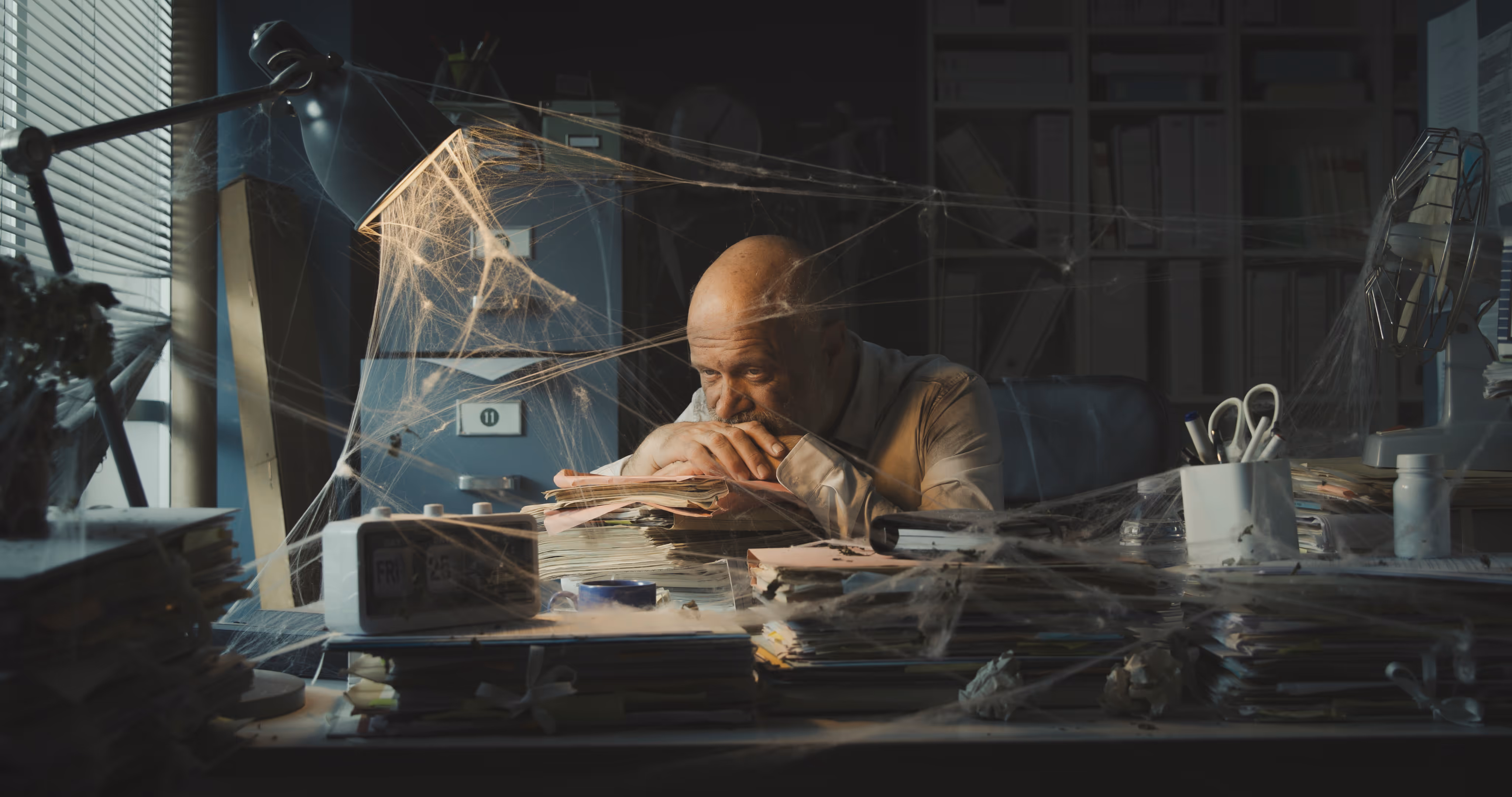 Mann sitzt traurig an einem mit Spinnweben bedeckten Schreibtisch voller Akten in einem düsteren Büro.
