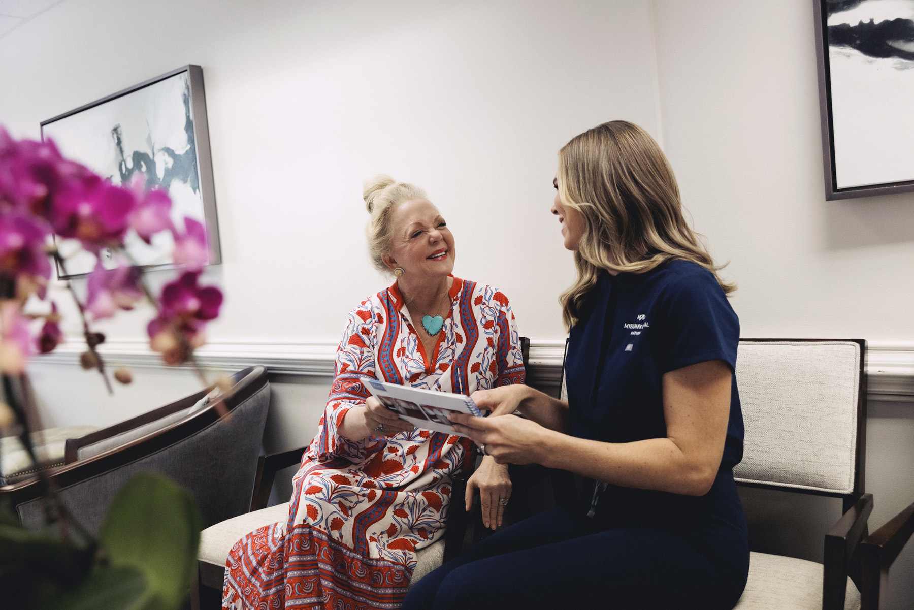 A senior woman in a colorful dress talking with a female medical professional in navy scrubs while holding a brochure in a waiting room.
