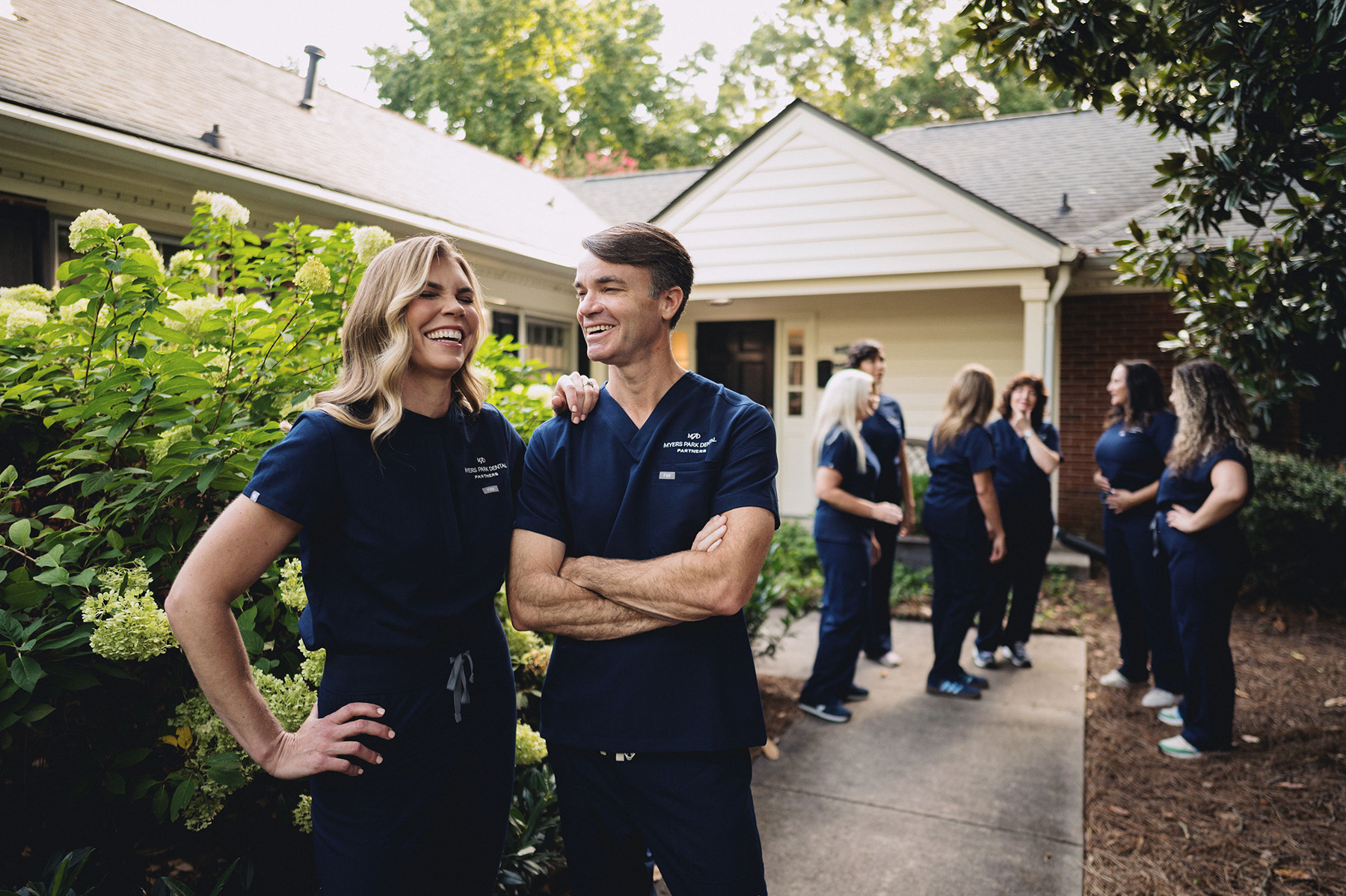 Two dental professionals in navy scrubs smiling and standing outdoors with a group of colleagues talking near a building in the background.