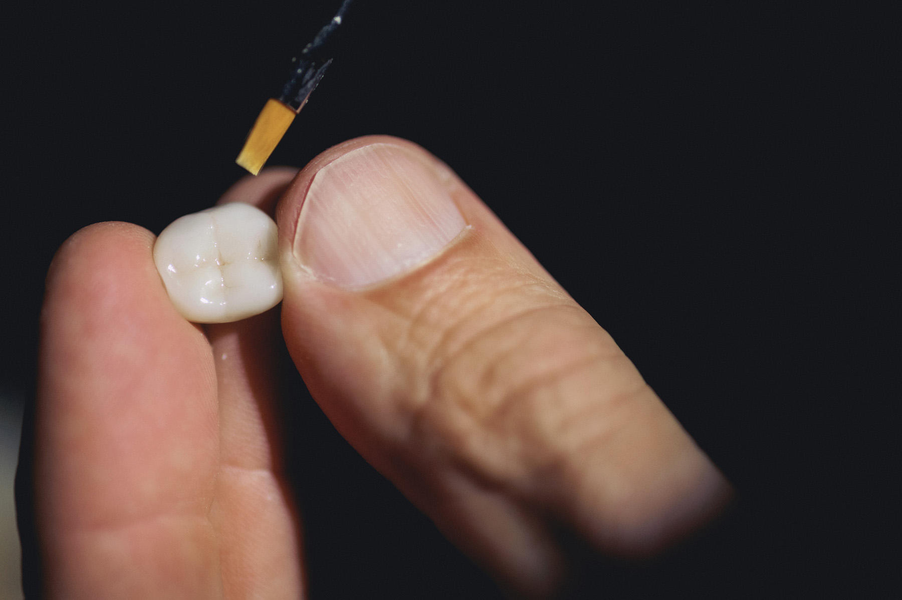 Hand holding a dental crown while a small brush applies a substance to it.