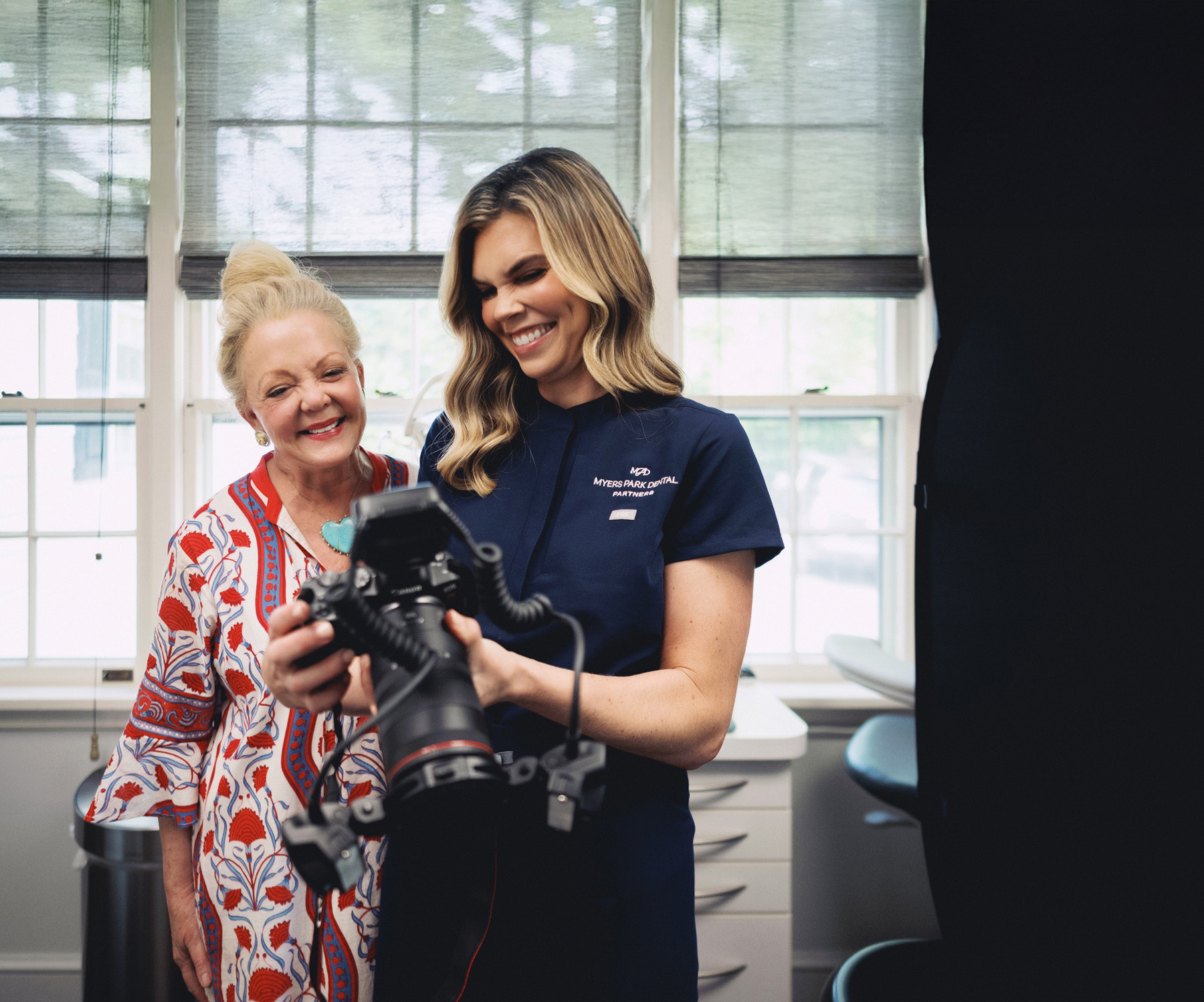 Two women smiling and looking at a digital camera in a bright room with large windows.