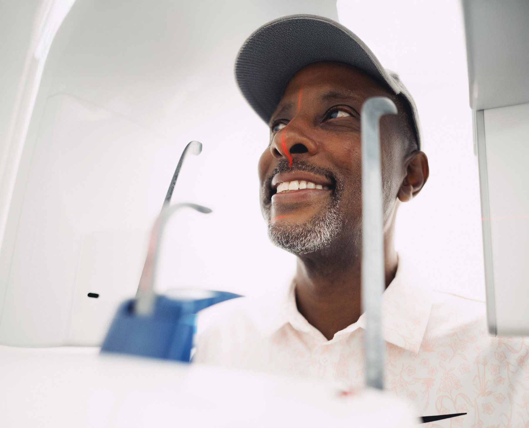 Smiling man wearing a cap undergoing a medical scan with red laser lines on his face.