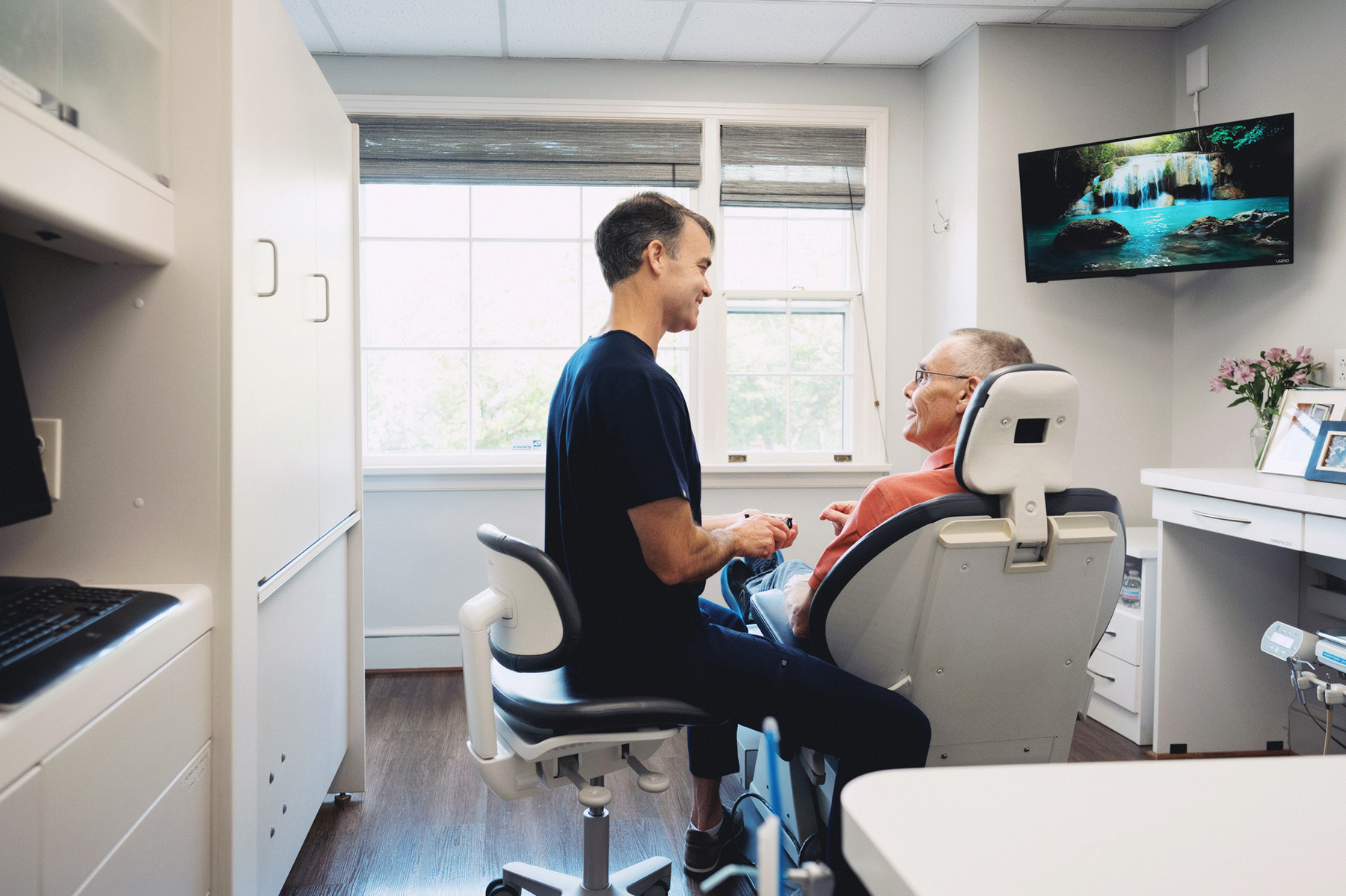 Dentist in navy scrubs smiling and talking with an older male patient seated in a dental chair in a bright clinic room.
