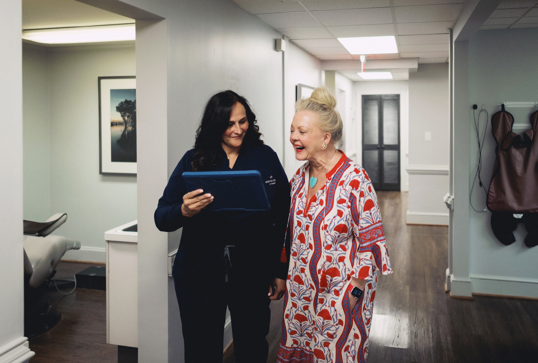 Two women in a medical office hallway looking at a tablet and smiling.
