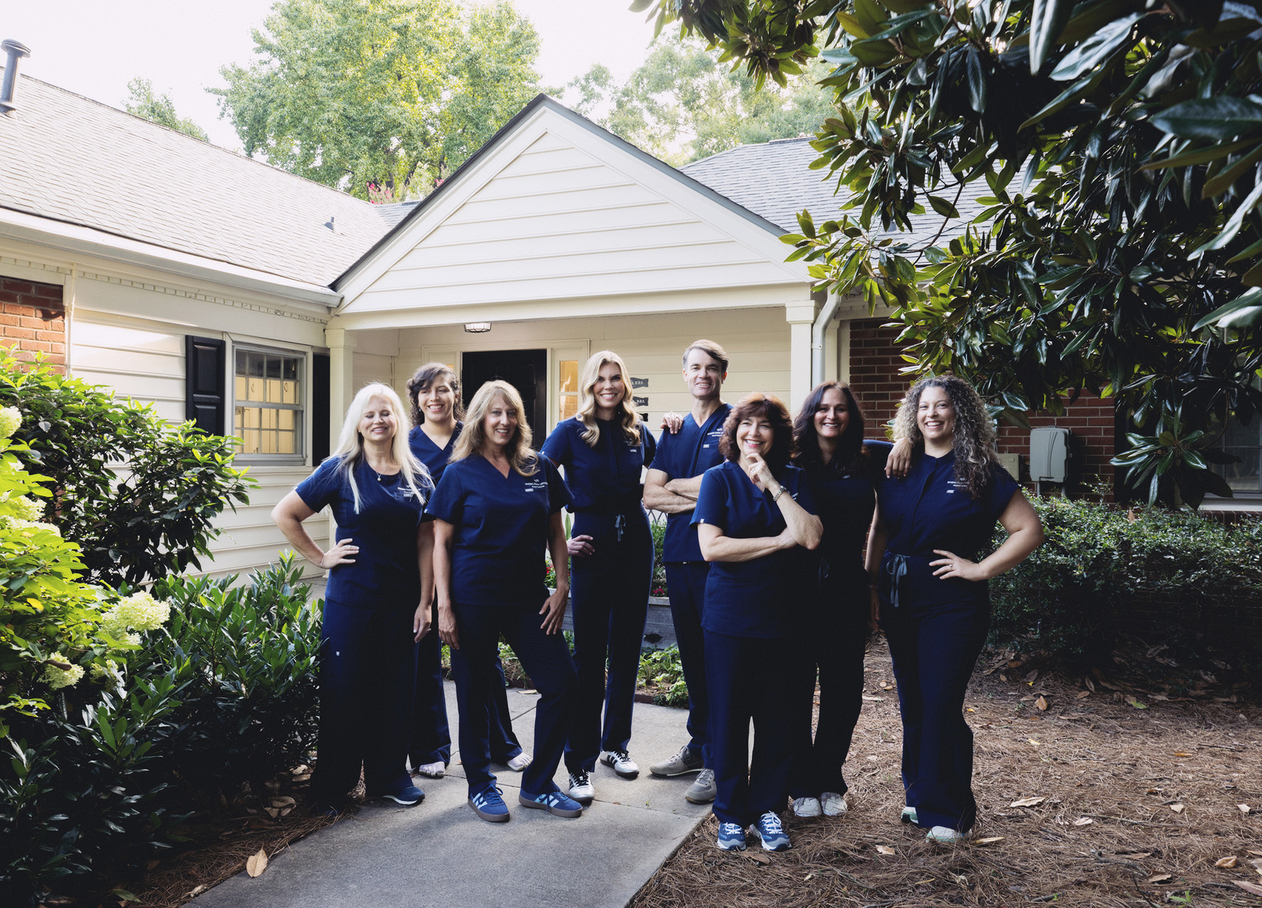 Group of eight healthcare professionals in navy scrubs standing outside a house with greenery and smiling at the camera.