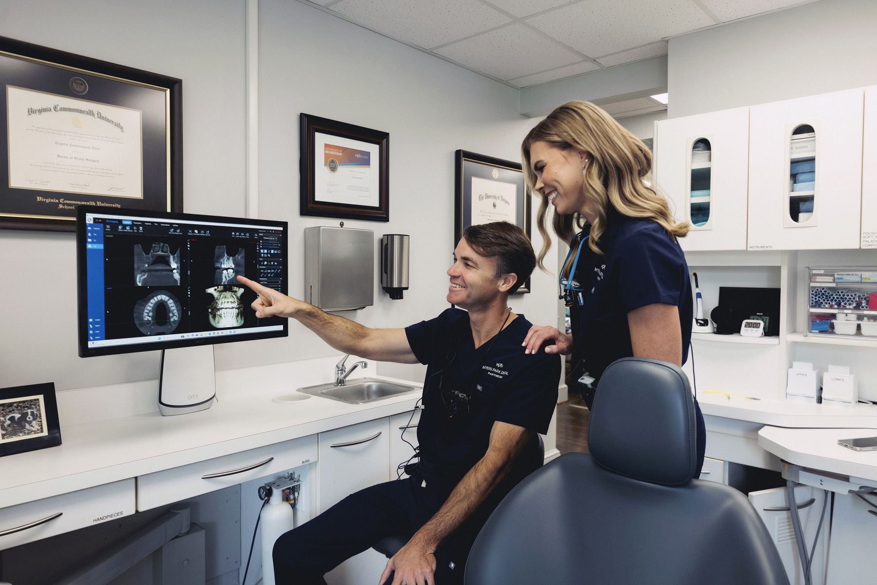 Two dental professionals in navy scrubs reviewing dental X-ray images on a computer screen in a clinic room.
