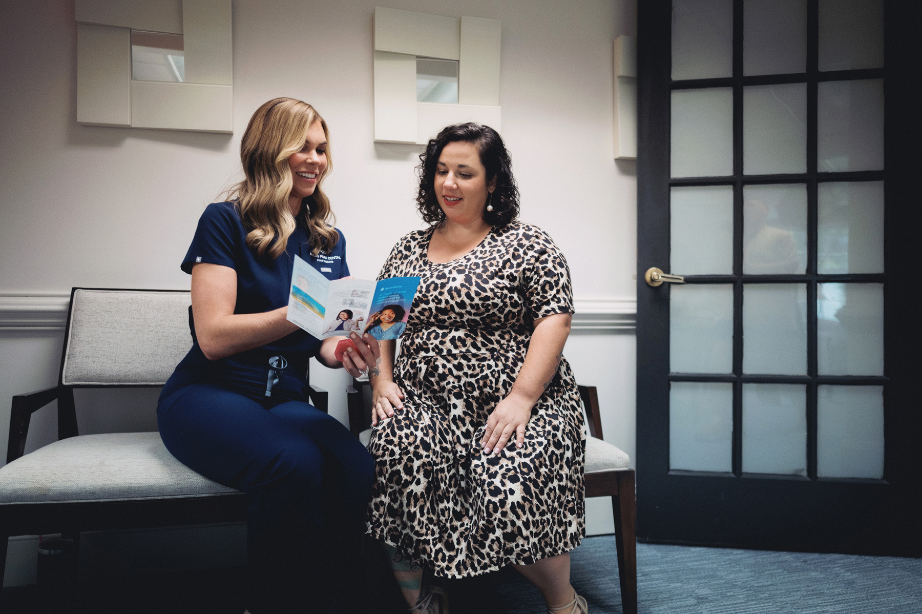 A female healthcare professional in navy scrubs shows a brochure to a woman in a leopard print dress sitting together on a bench.