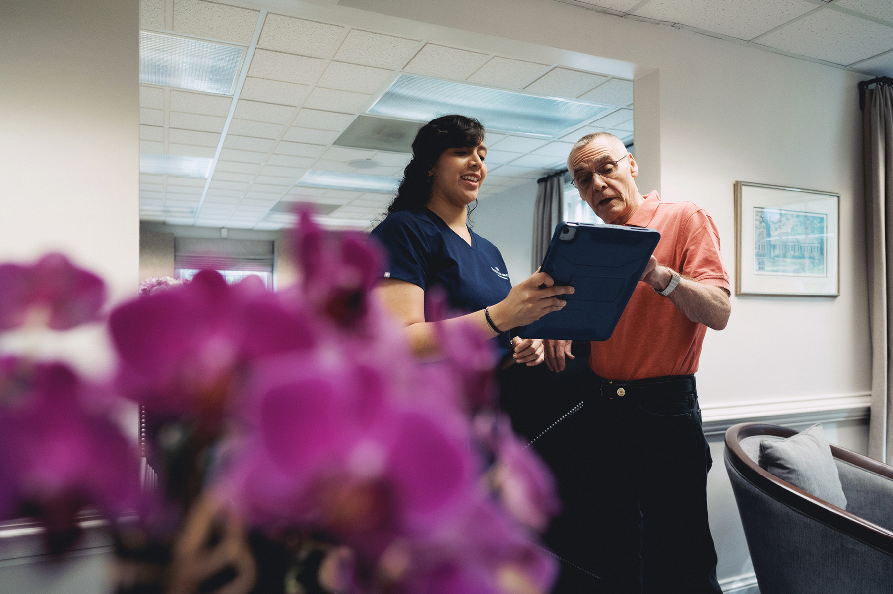 Healthcare professional in navy scrubs shows a tablet to an older man in a coral shirt inside a room with purple flowers in the foreground.