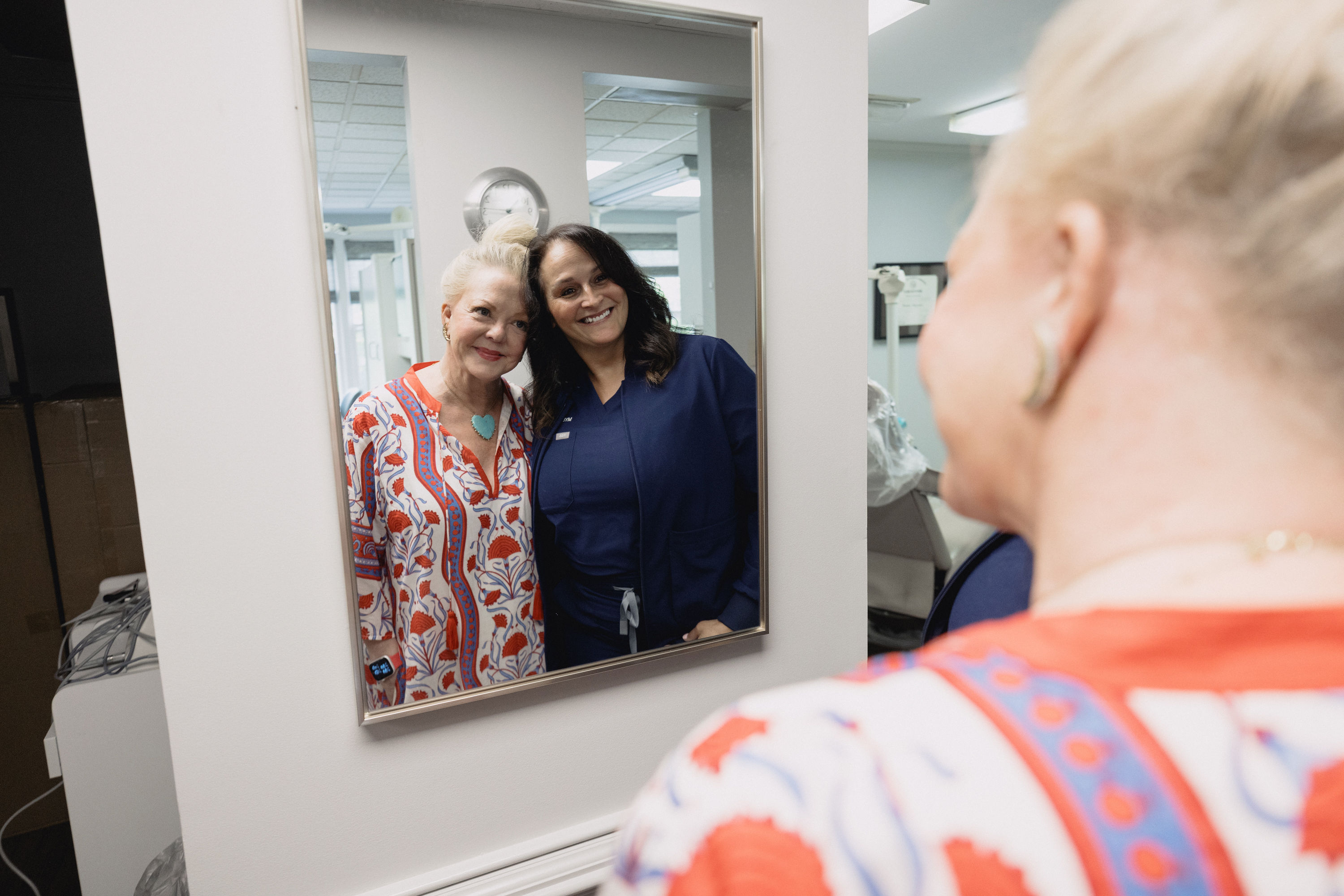 Two women smiling and posing closely in front of a mirror inside a clinic or office.