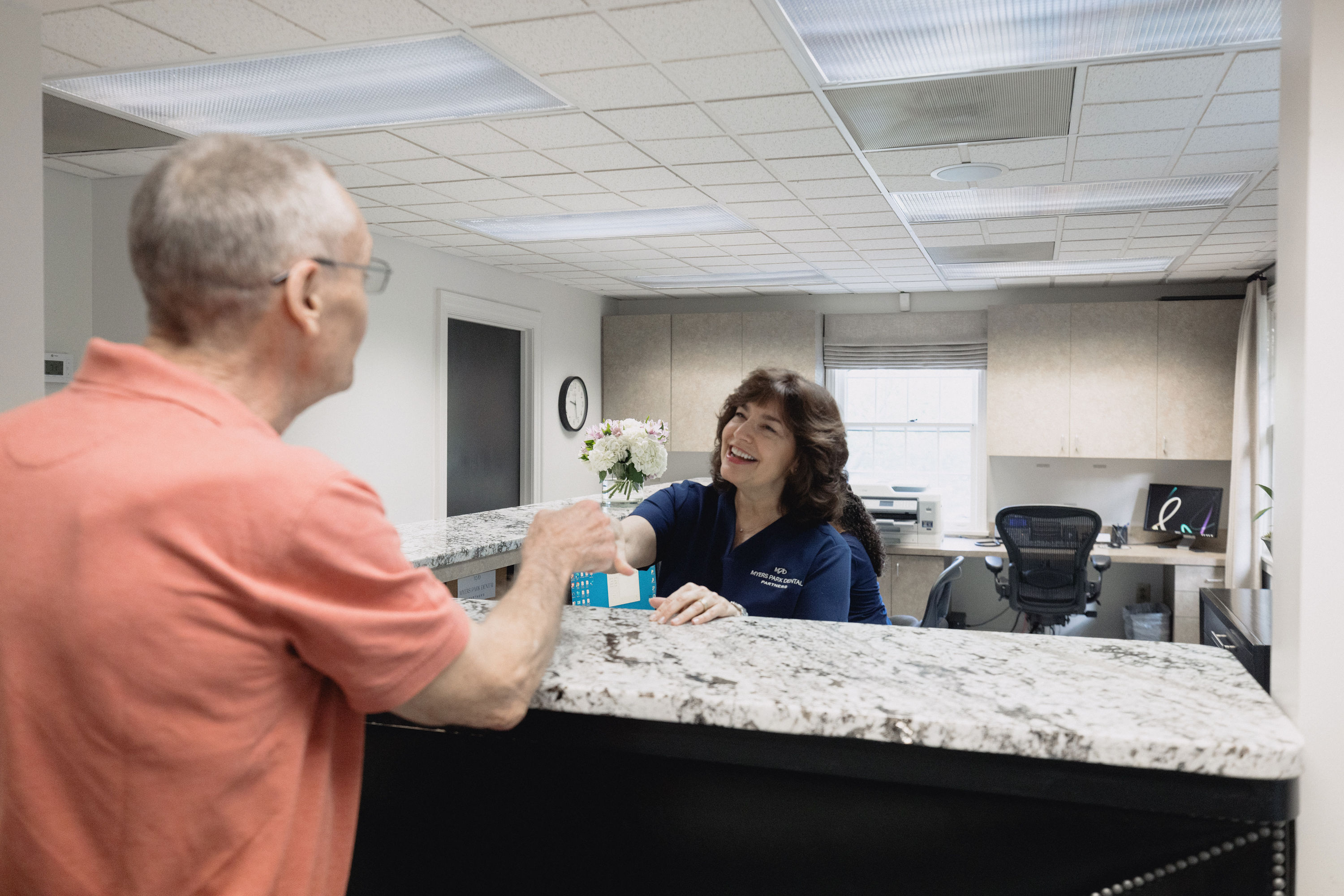 Receptionist in navy uniform smiling and handing a card to an elderly man at a dental office front desk.