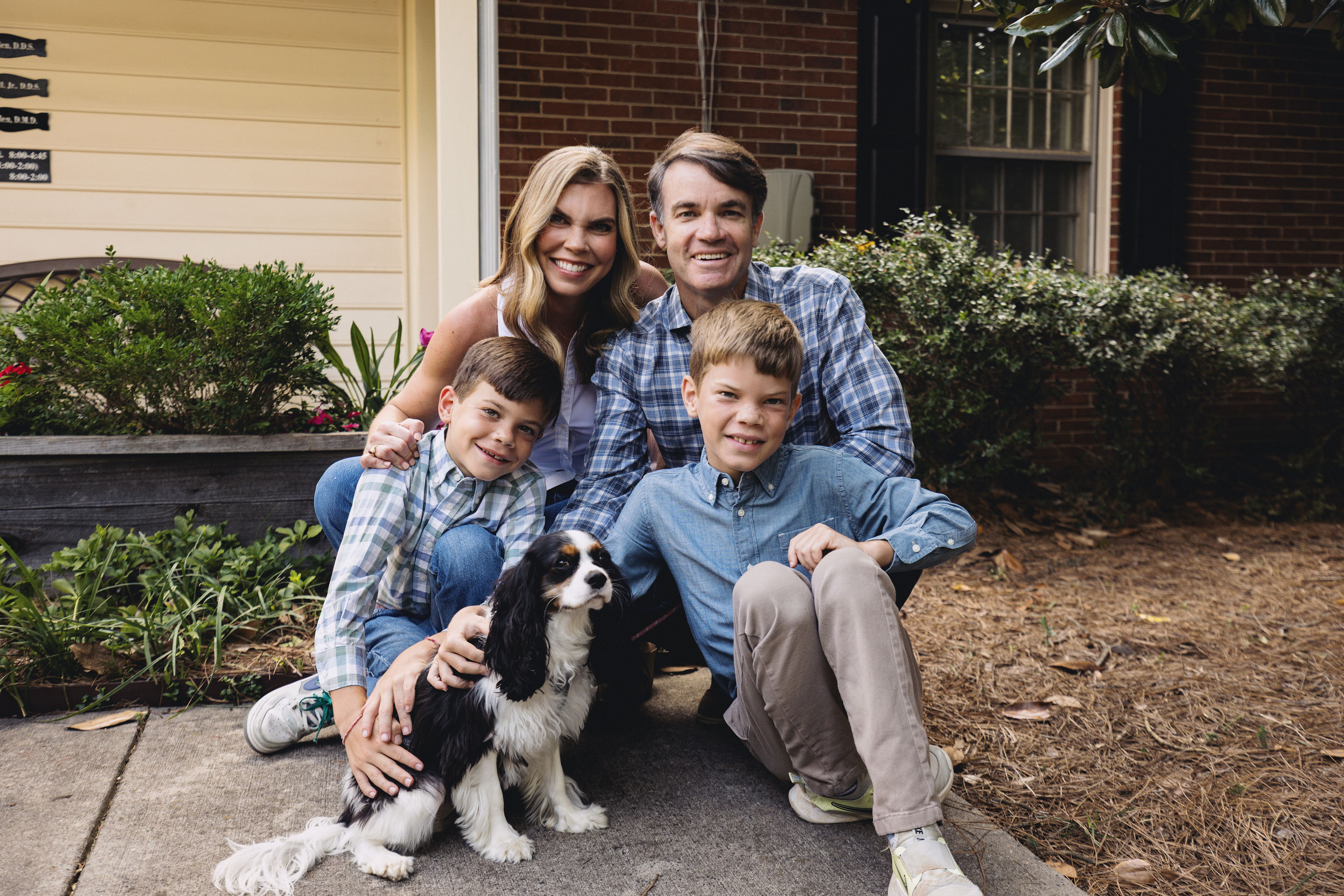 Smiling family of four, with mother, father, two boys, and a black and white dog, posing outside a suburban house.